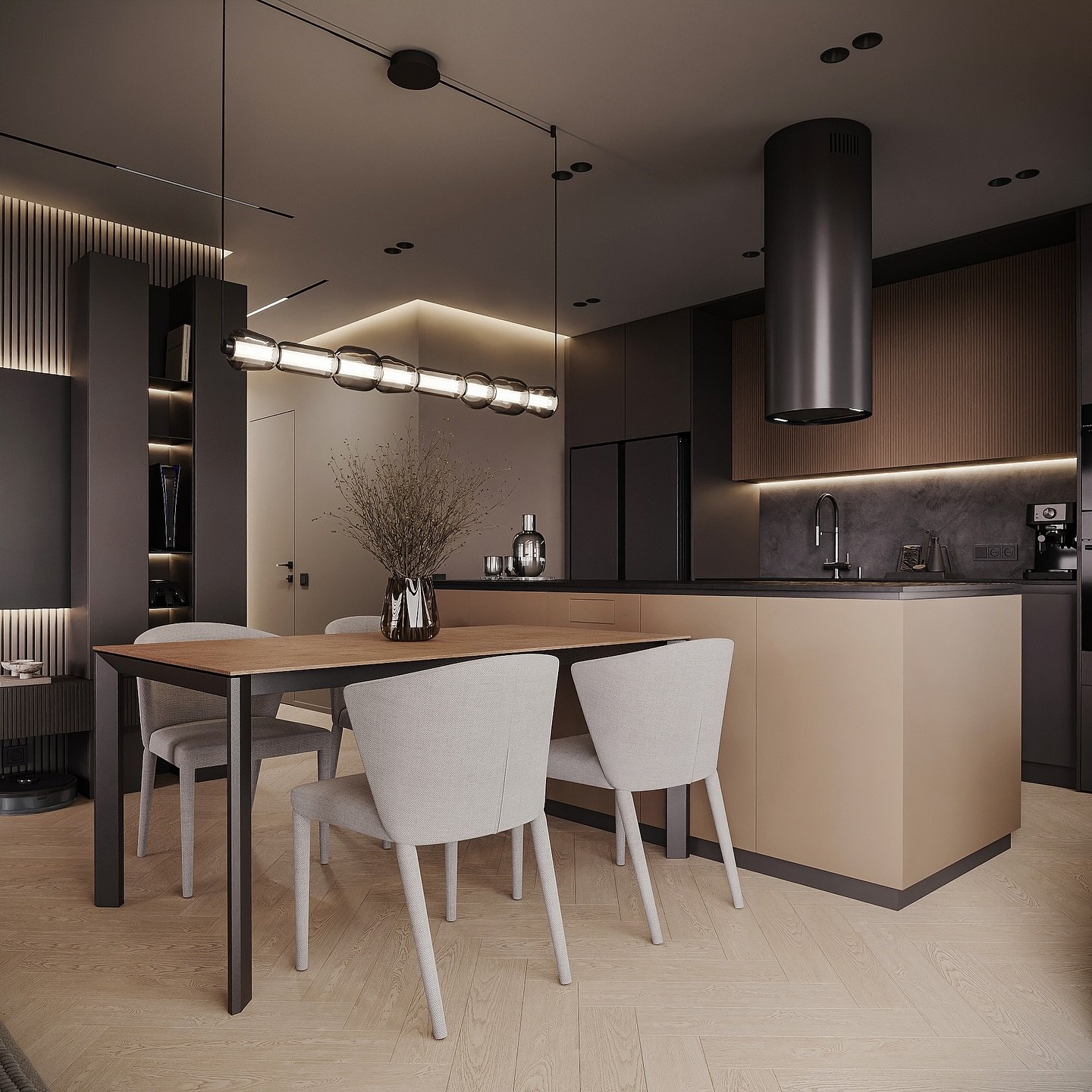 A dark and sophisticated open-concept studio space. The kitchen features dark grey and wood cabinetry with a beige island. In the foreground is a dining table with a light wood top and four grey chairs. A long, multi-bulb pendant light hangs over the dining area.