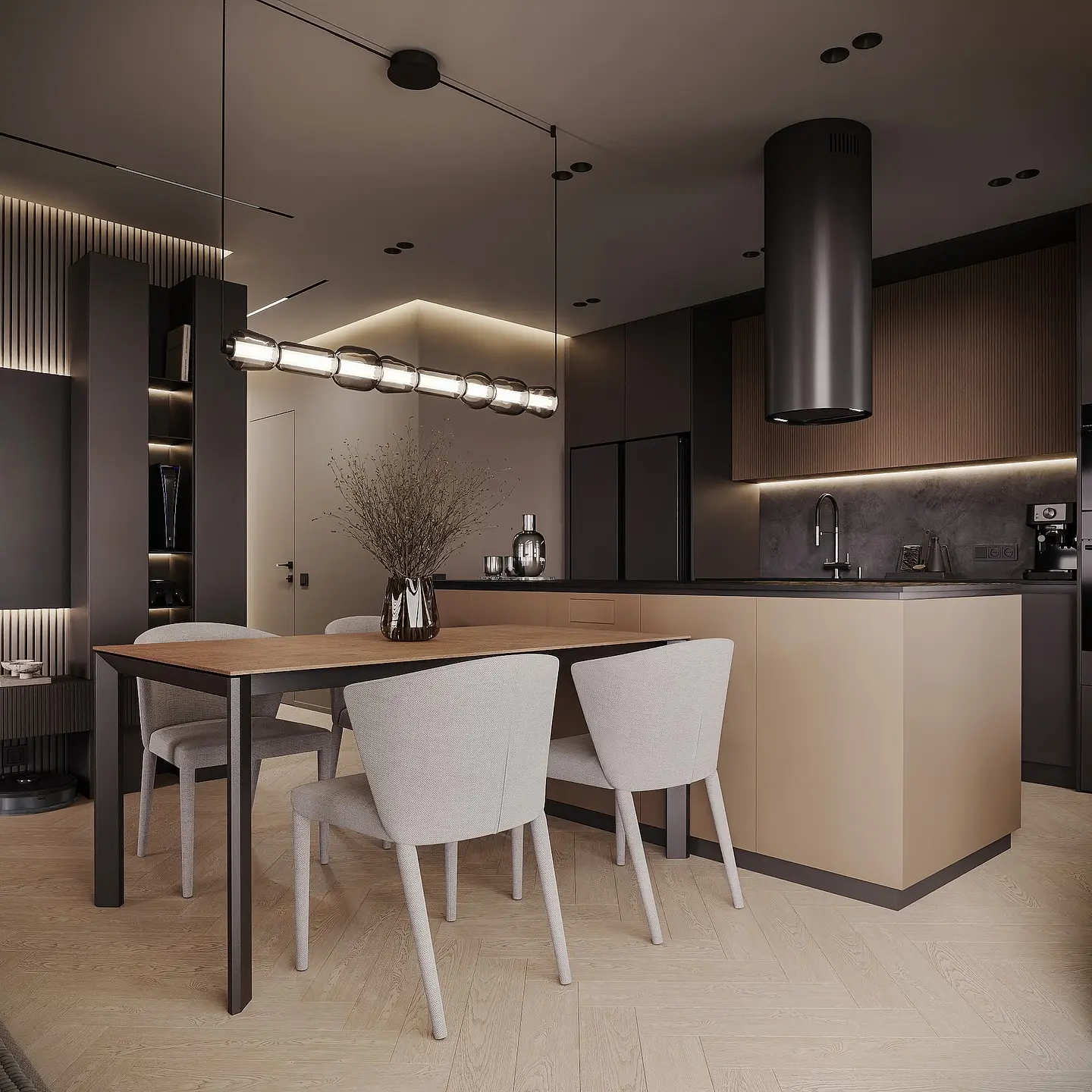 A dark and sophisticated open-concept studio space. The kitchen features dark grey and wood cabinetry with a beige island. In the foreground is a dining table with a light wood top and four grey chairs. A long, multi-bulb pendant light hangs over the dining area.
