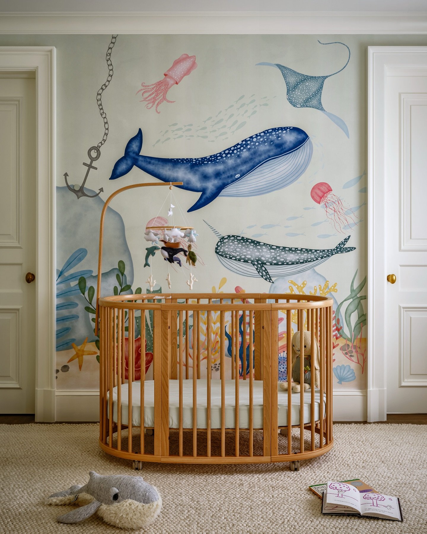 A nursery with a light wood oval crib and a large watercolor-style mural featuring whales, manta rays, and jellyfish.
