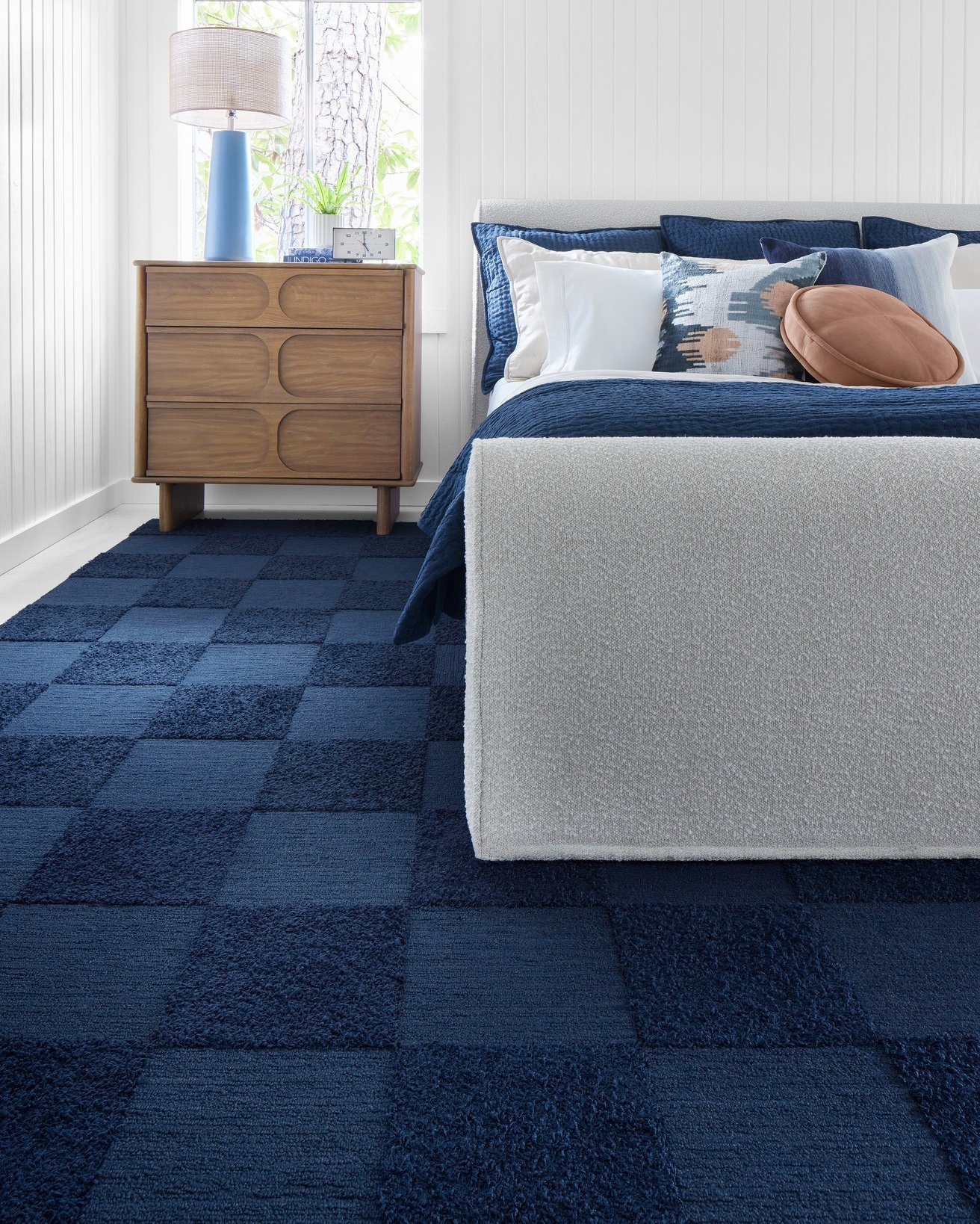 A large, checkerboard-patterned rug made of alternating squares of two shades of deep blue, placed under a bed with blue and white bedding.