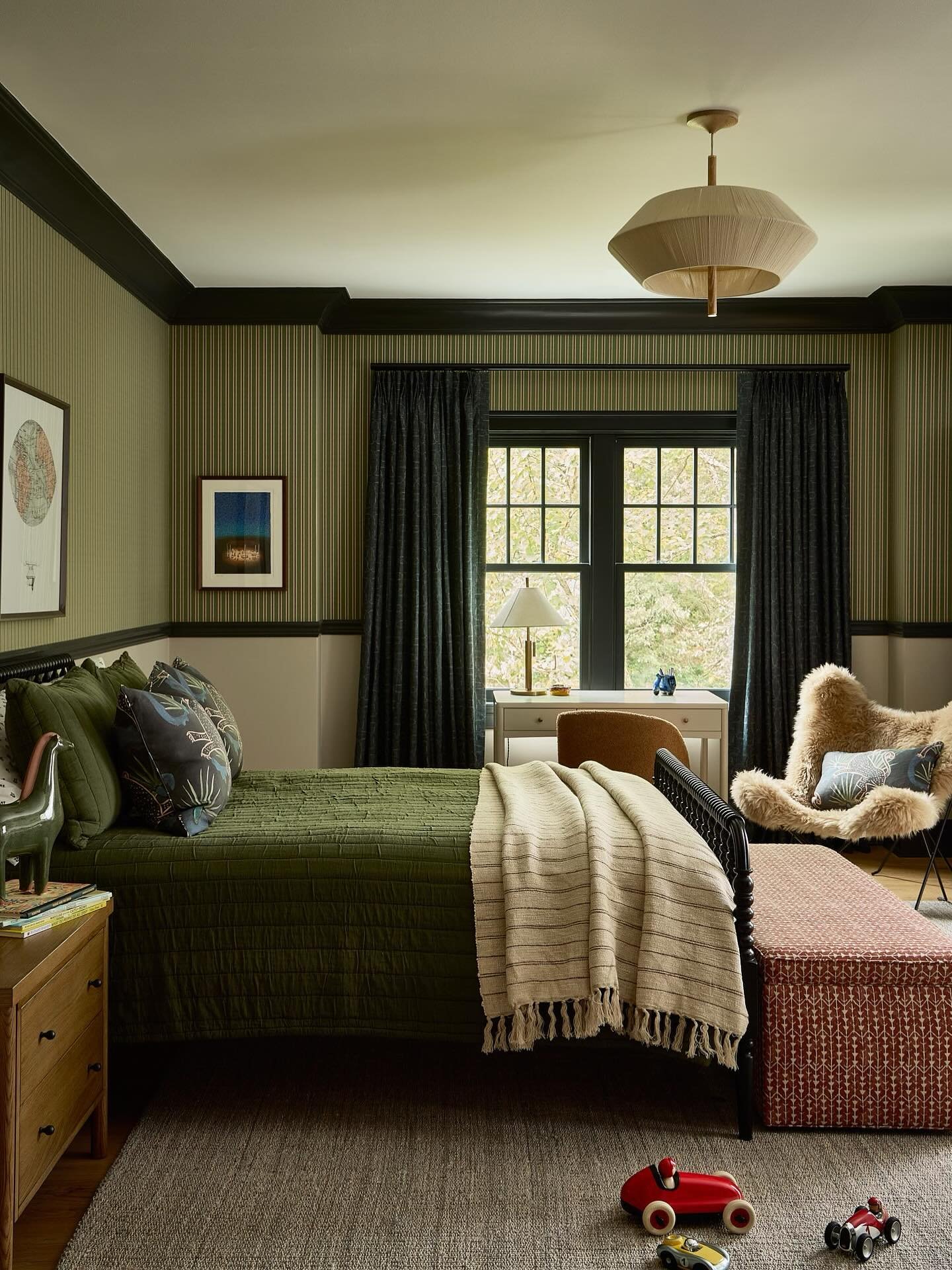 A dark, moody bedroom with green and striped wallpaper, a daybed, and vintage decor, including a toy dinosaur and a wooden car.