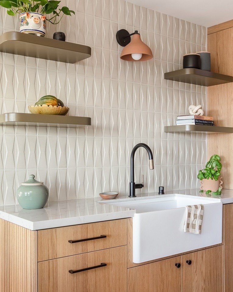 A kitchen featuring a white, three-dimensional geometric tile backsplash, paired with a farmhouse sink, light wood cabinetry, and sage green floating shelves.