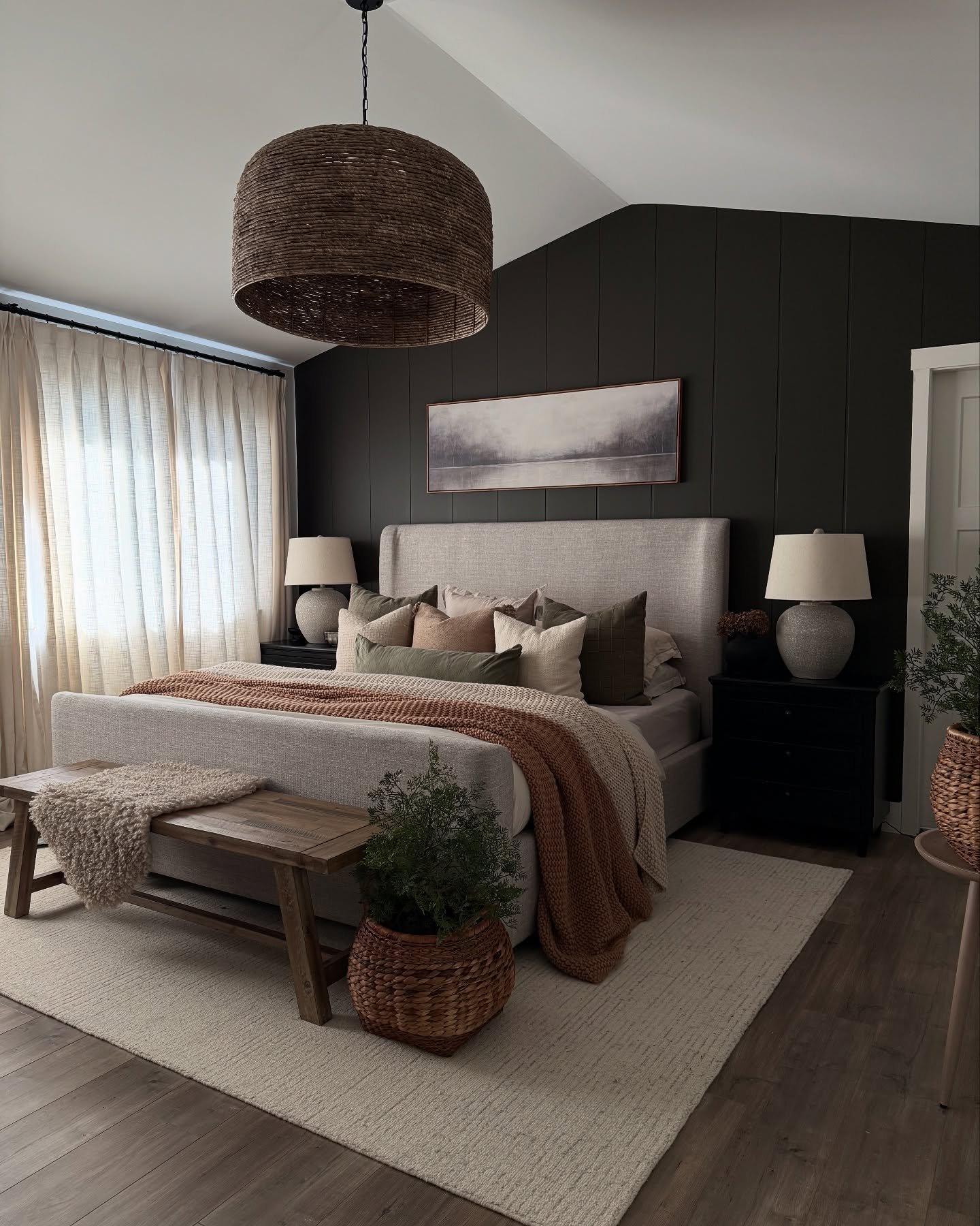 A cozy bedroom with a dark accent wall, a light-colored upholstered bed, a large woven pendant light, and a wooden bench at the foot of the bed.