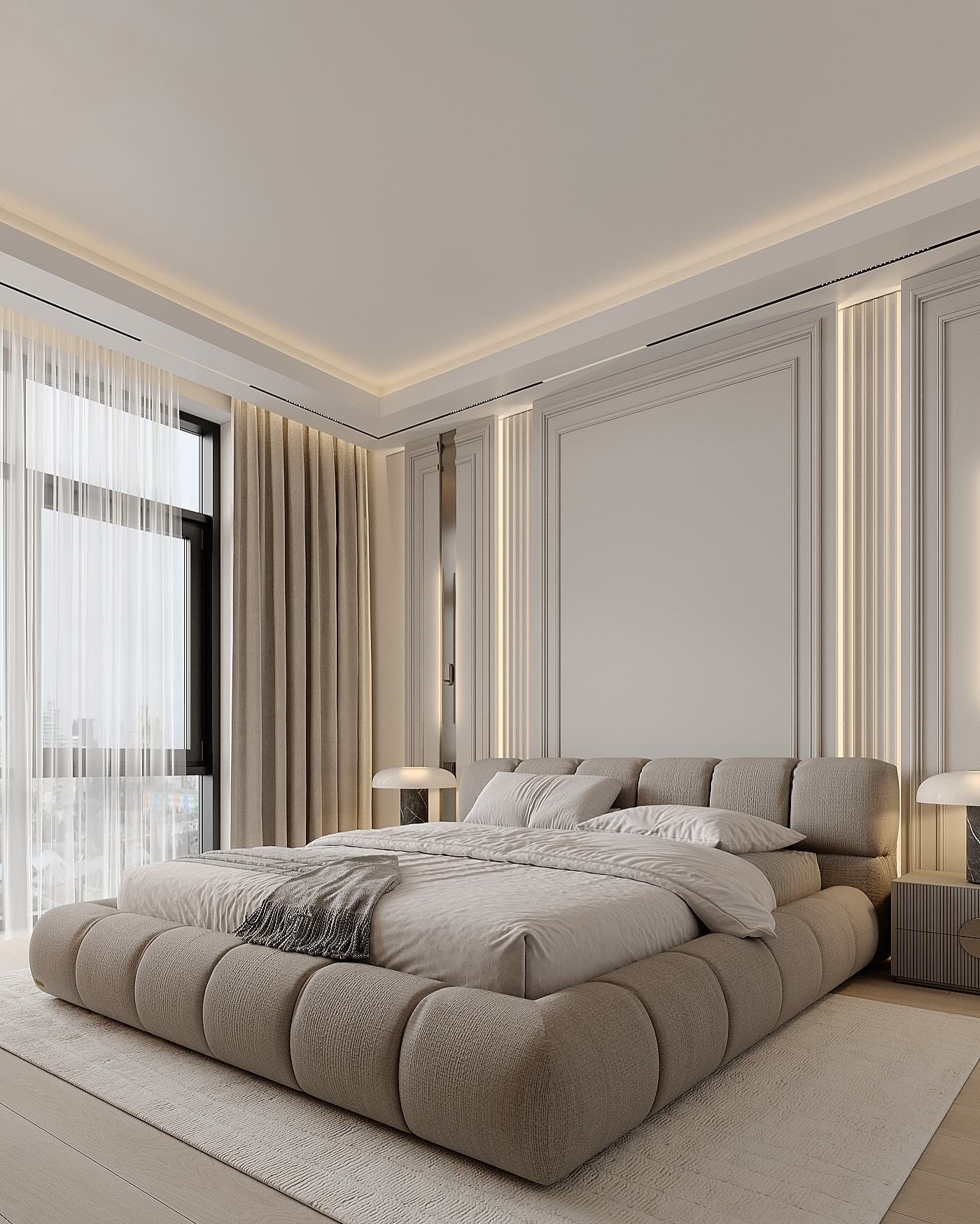 A luxurious bedroom with a large, low-profile upholstered bed featuring a unique quilted, puffy design. The room has a neutral color palette, tall windows, and a headboard wall with classic molding and vertical light strips.