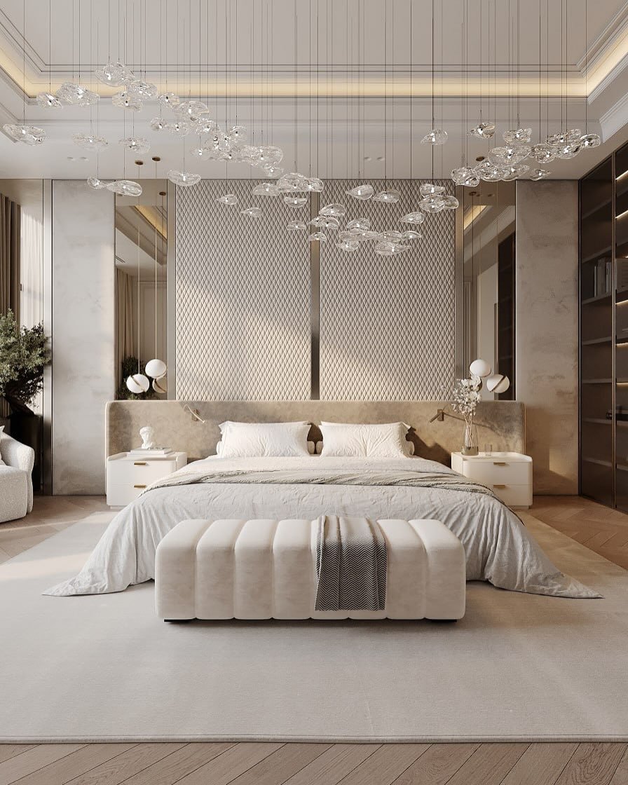 A luxurious and bright master bedroom with a large, tufted bench at the foot of the bed. The room features a stunning, cloud-like chandelier made of multiple clear, sculptural light fixtures.