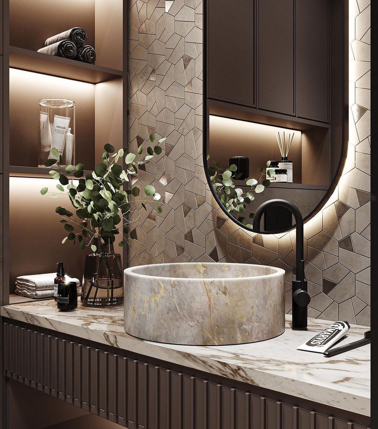 A luxurious bathroom featuring a cylindrical marble vessel sink with gold veining, set on a matching marble countertop. A black faucet and a unique geometric tiled wall complete the modern, high-end look.