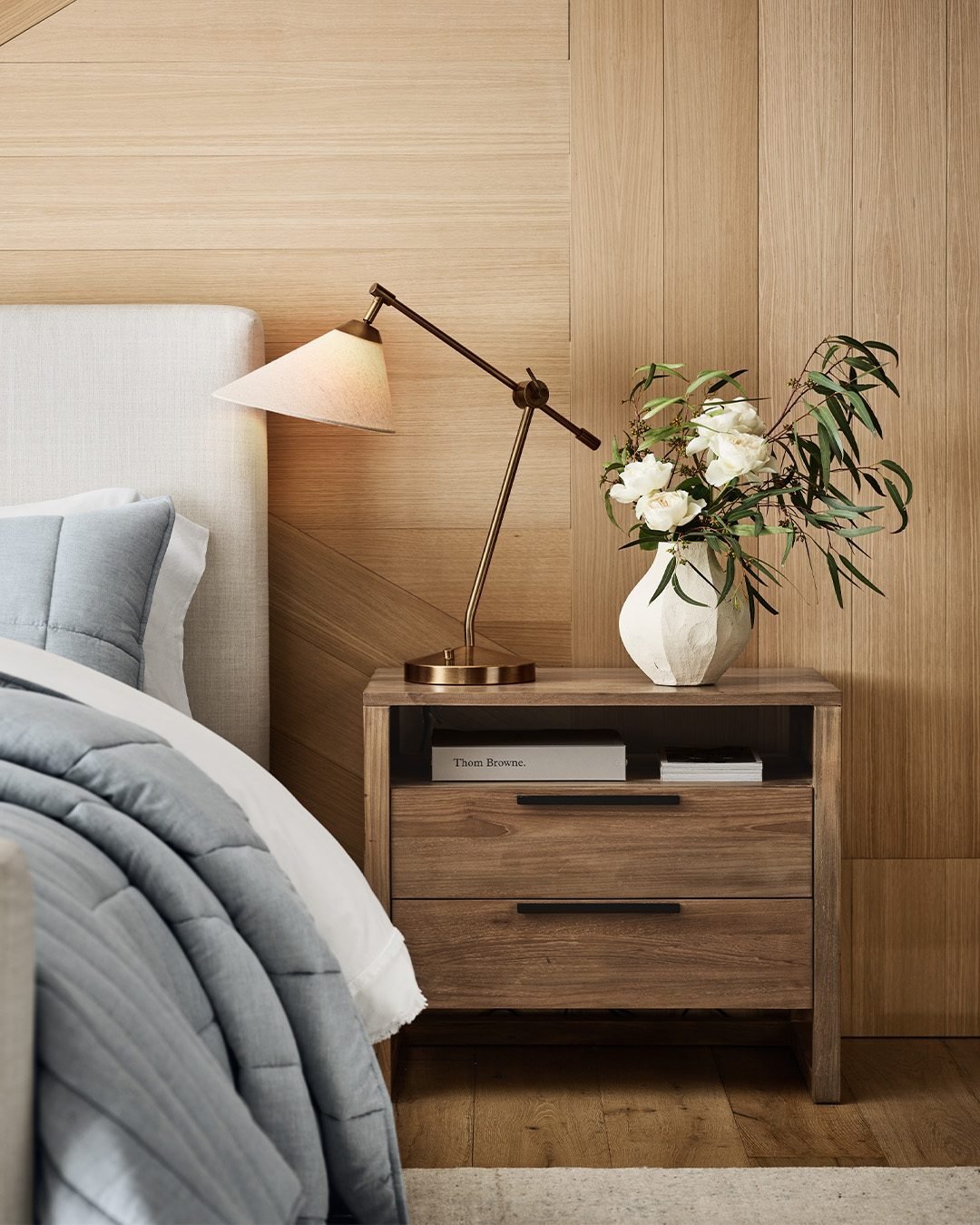 A close-up shot of a rustic wooden nightstand with two drawers and an open shelf. It holds a modern brass lamp with a white glass shade and a white vase with eucalyptus and white flowers.