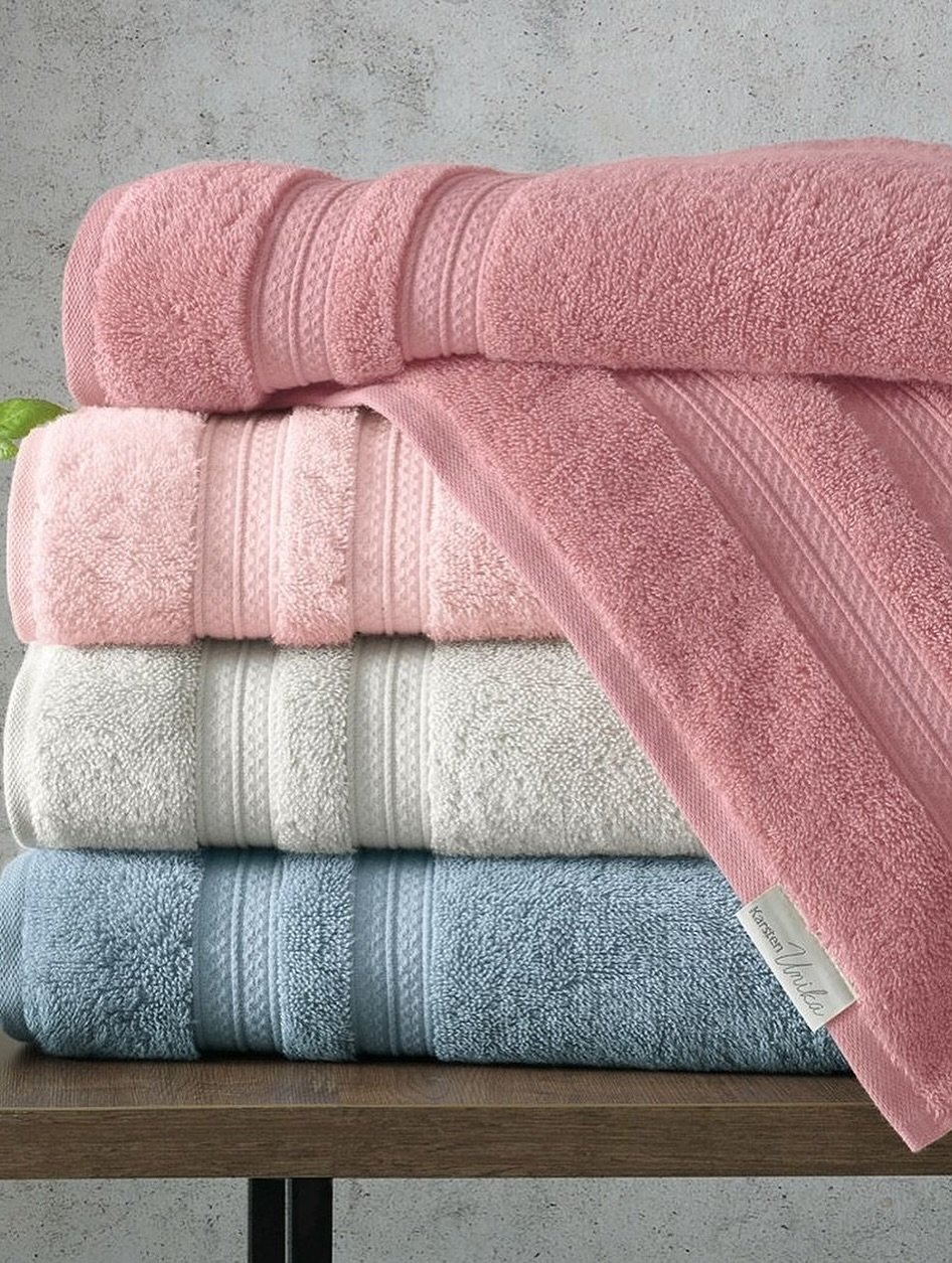 Stack of four plush bath towels in dusty rose, blush pink, white, and powder blue on a wooden shelf.