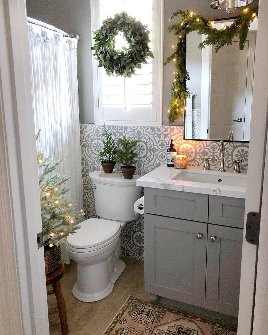 A neutral grey and white farmhouse bathroom decorated for Christmas with a wreath, lighted mirror garland, and small potted trees.