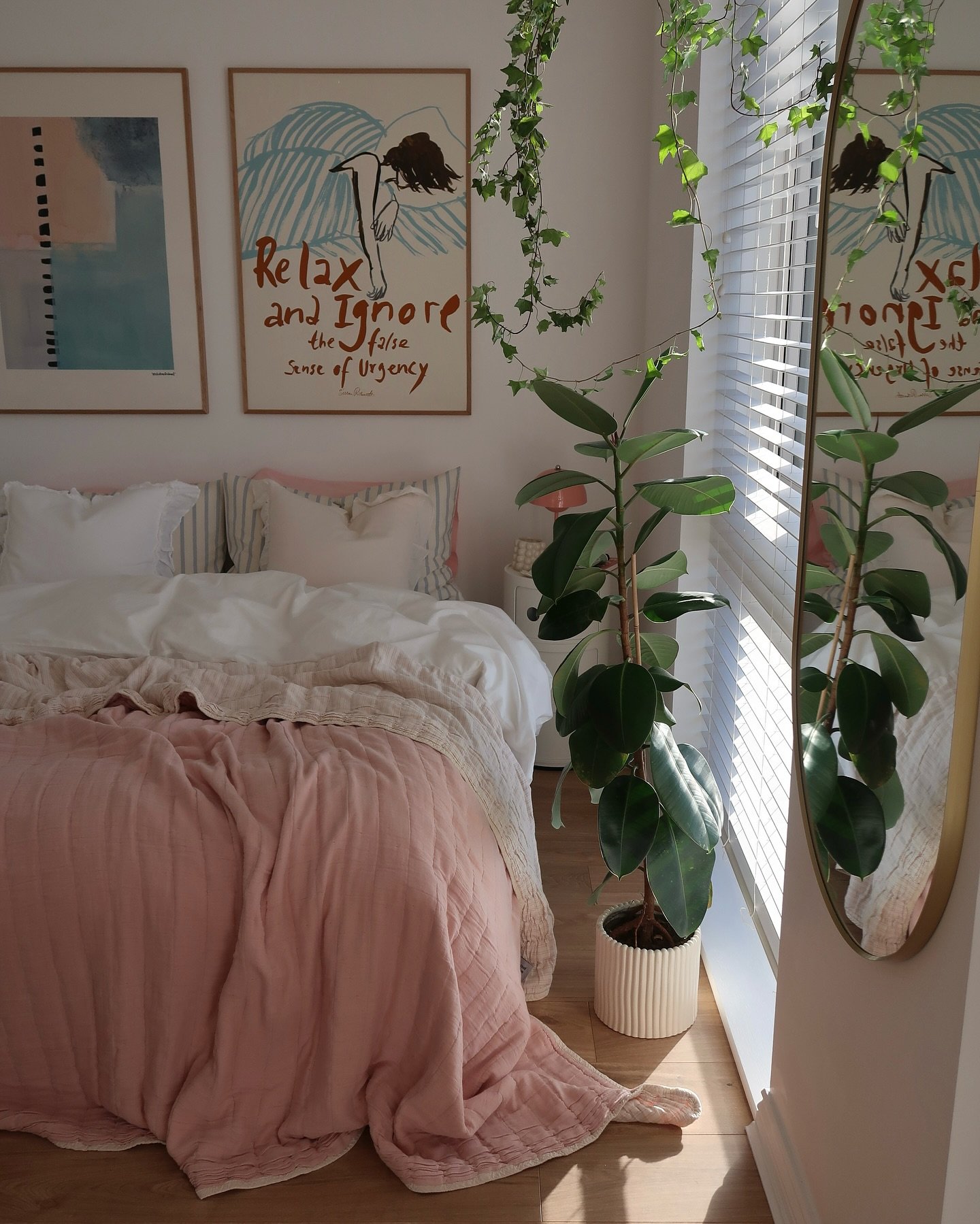 A cozy bedroom featuring a bed with a light-pink duvet, a large potted plant, and a gold-framed mirror reflecting the sunny window.