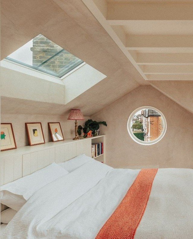 A bright loft bedroom with a large skylight and a small round window. The room features warm, light-colored walls, a bed with white and terracotta-colored bedding, and a built-in headboard.