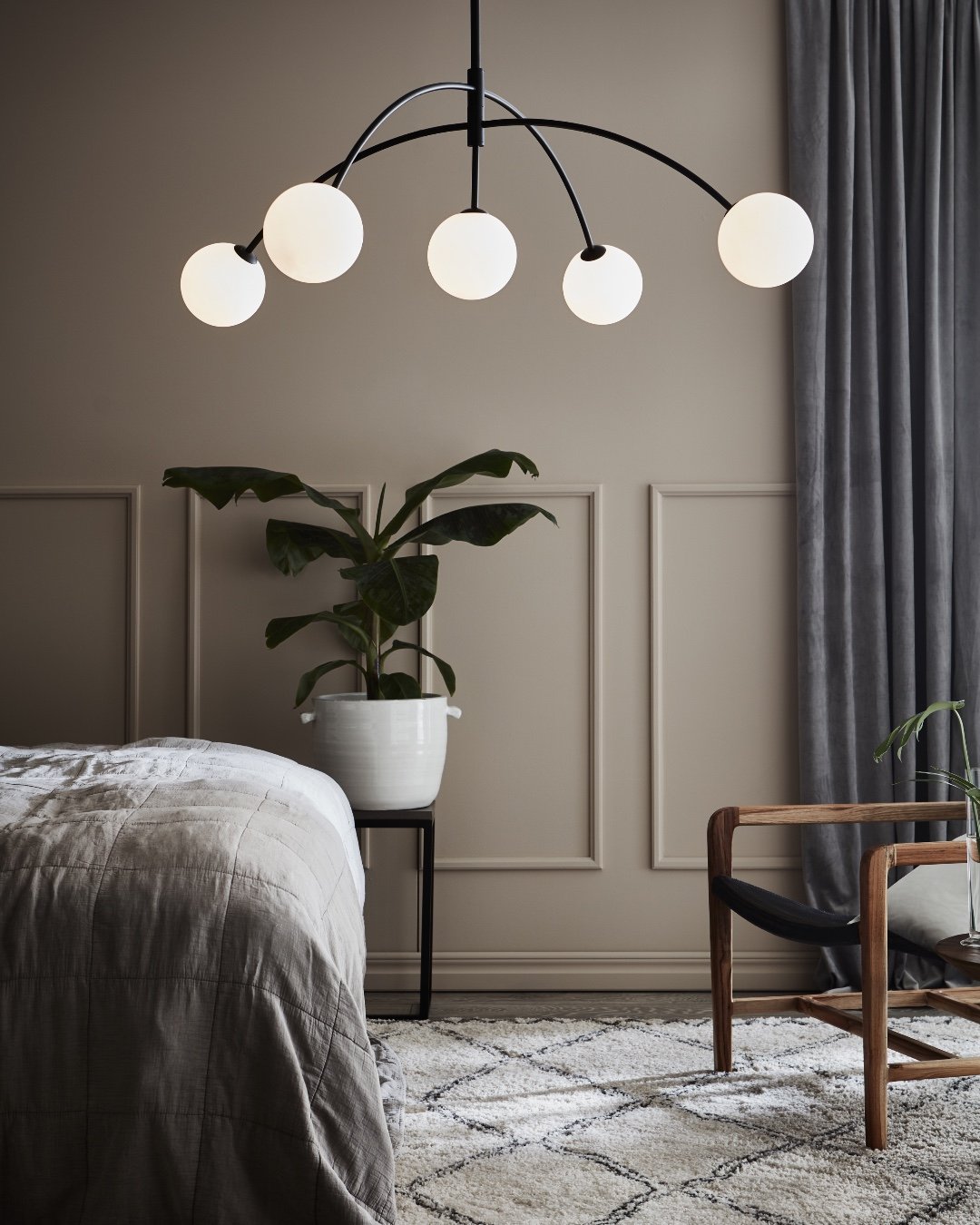 A contemporary black chandelier with five white orb-shaped lights hangs over a bed in a minimalist bedroom.