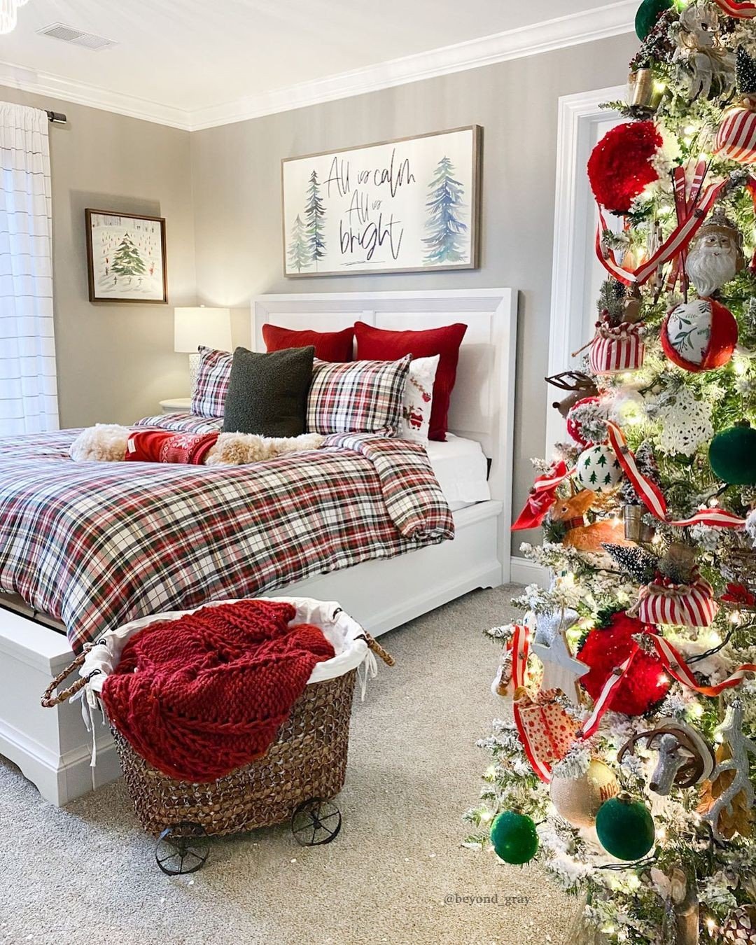 A bedroom decorated for Christmas featuring a bed with red and white plaid bedding, a large flocked Christmas tree with red and green ornaments, and a woven basket with a red knit blanket.