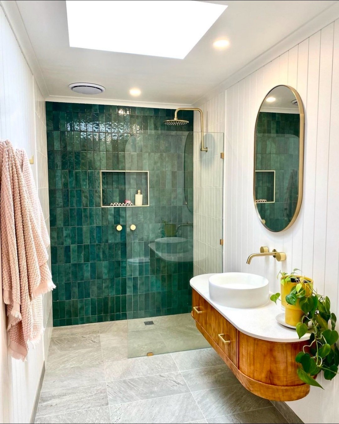 A modern bathroom with a dark emerald green tiled shower wall, white vertical shiplap walls, a floating wooden vanity, and gold fixtures.