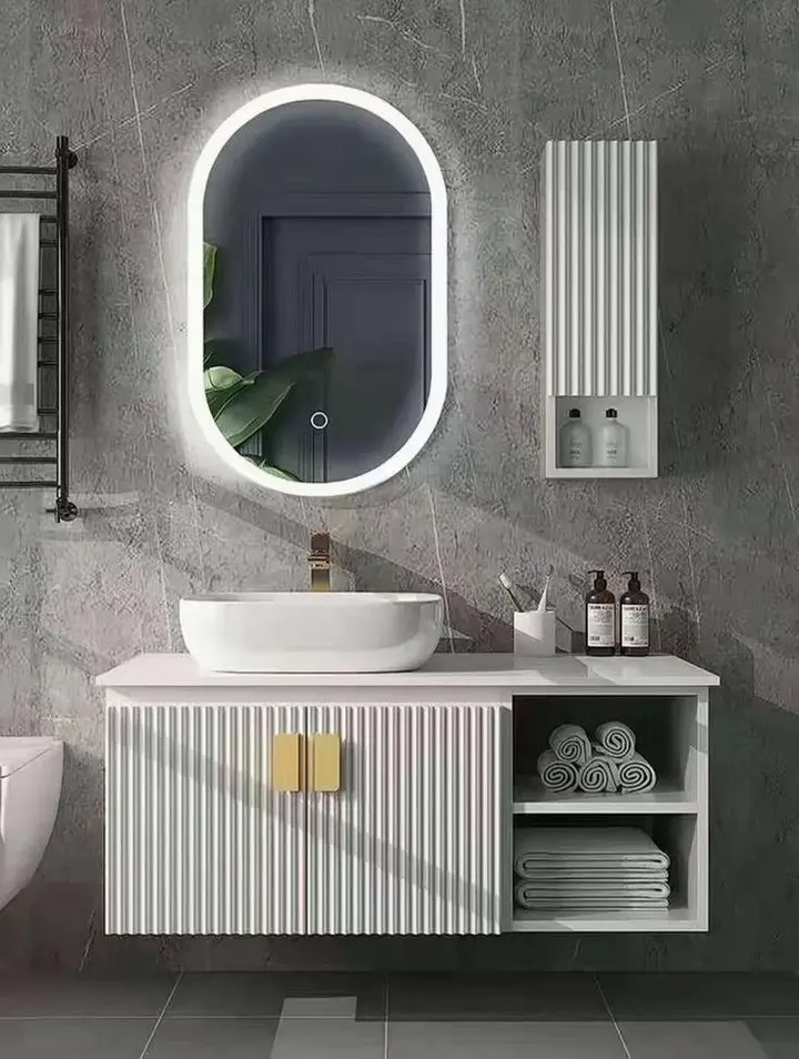 A white floating bathroom vanity with a ribbed wood texture and gold handles, paired with a vessel sink, an oval backlit mirror, and a matching wall cabinet, against a dark gray tiled wall.