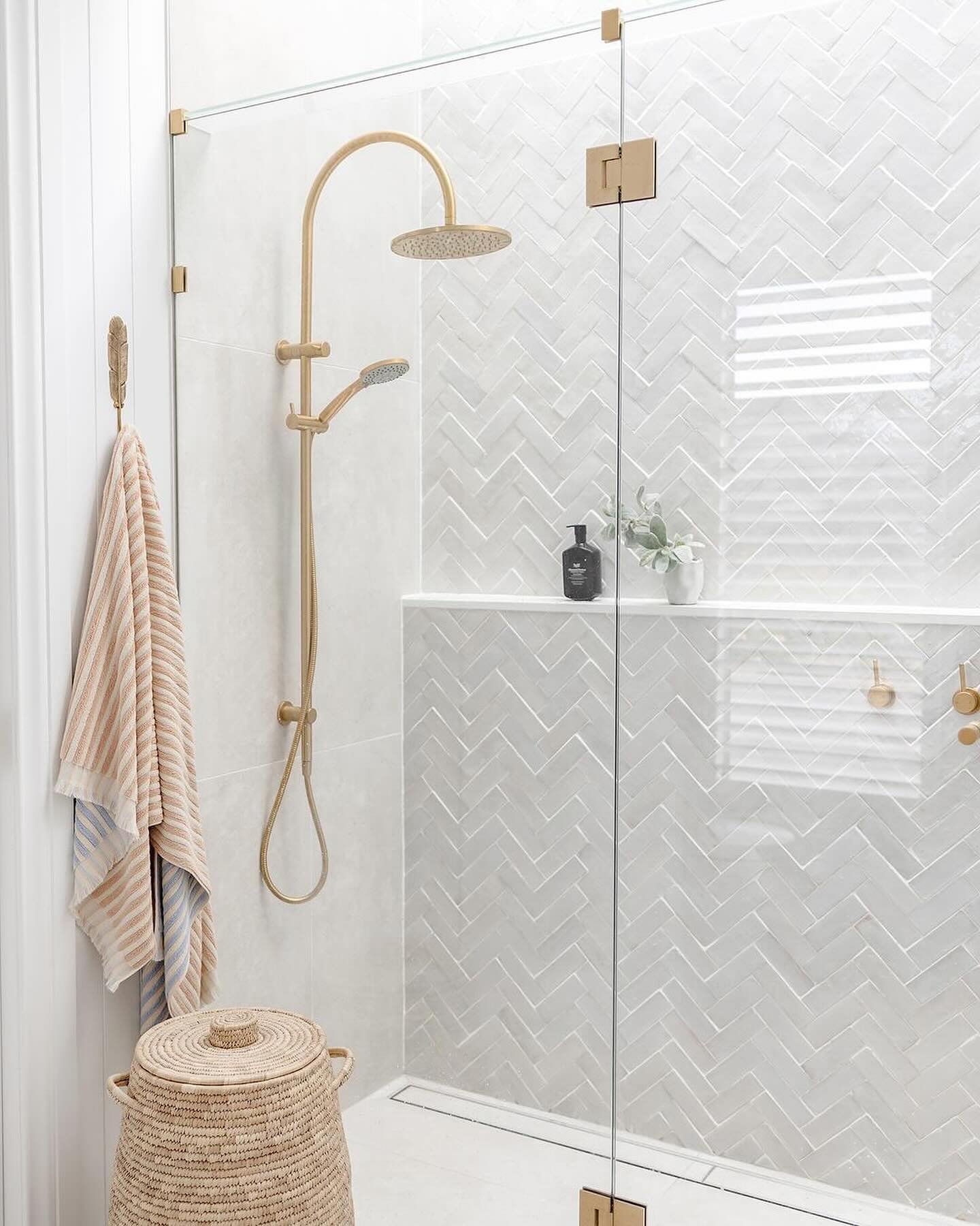 A bright, modern shower featuring white glazed porcelain tiles in a herringbone pattern with contrasting pale gray grout and brushed gold fixtures.