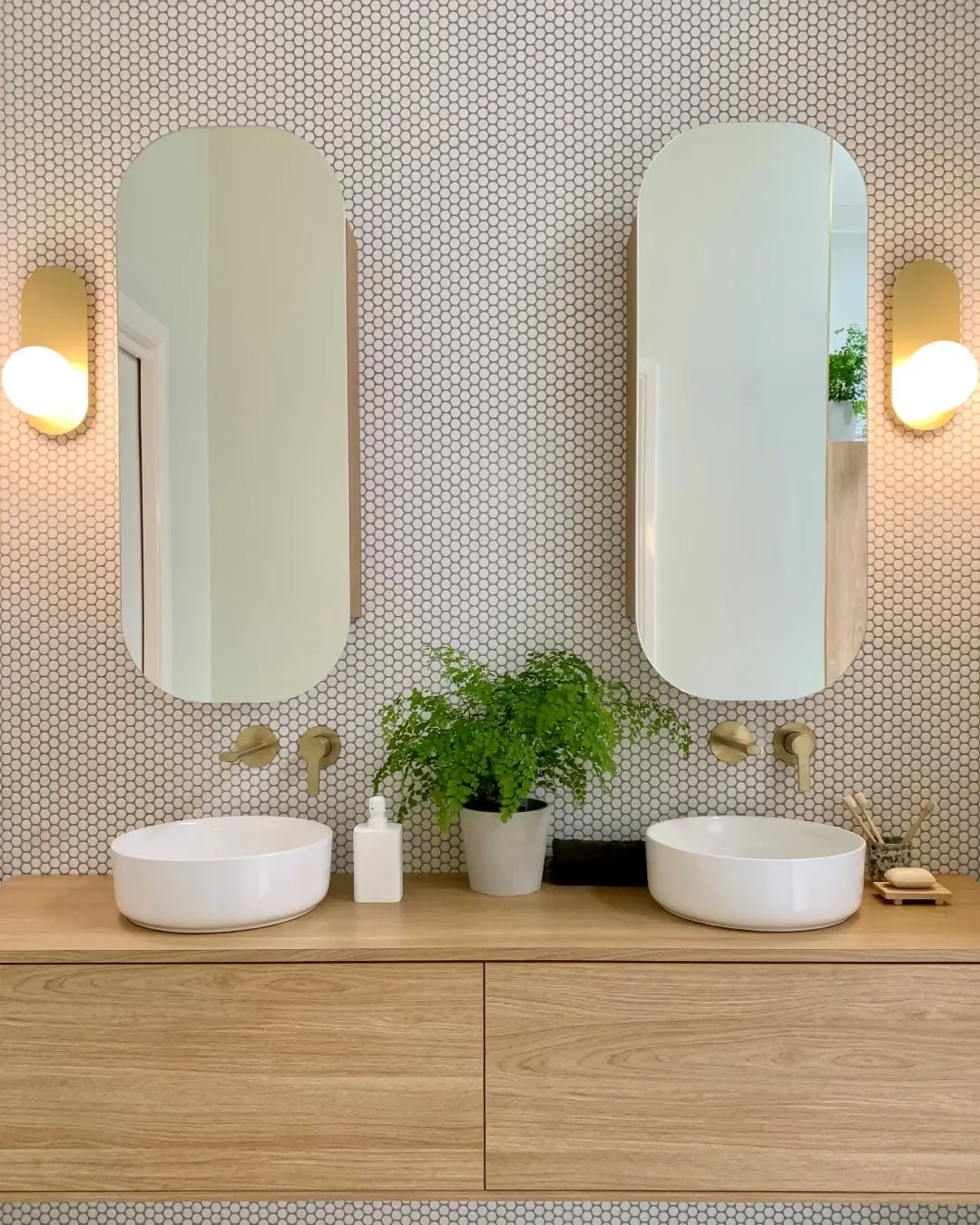 A bright bathroom with a double sink vanity and two identical pill-shaped mirrors with thin gold frames against a white hexagonal tiled wall.