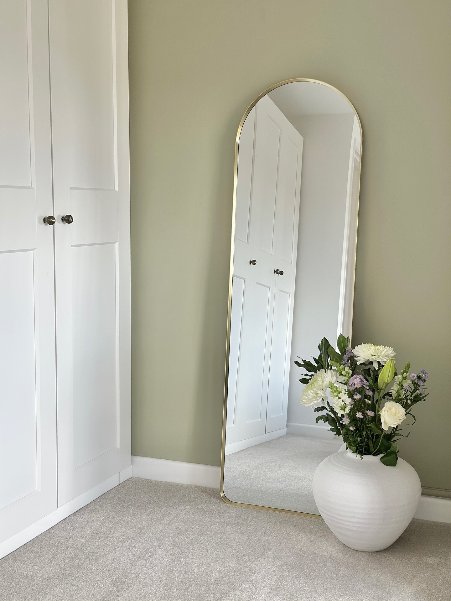 A tall, slender gold-framed arch mirror stands in a bedroom corner, next to a large white vase filled with flowers, reflecting white wardrobes.