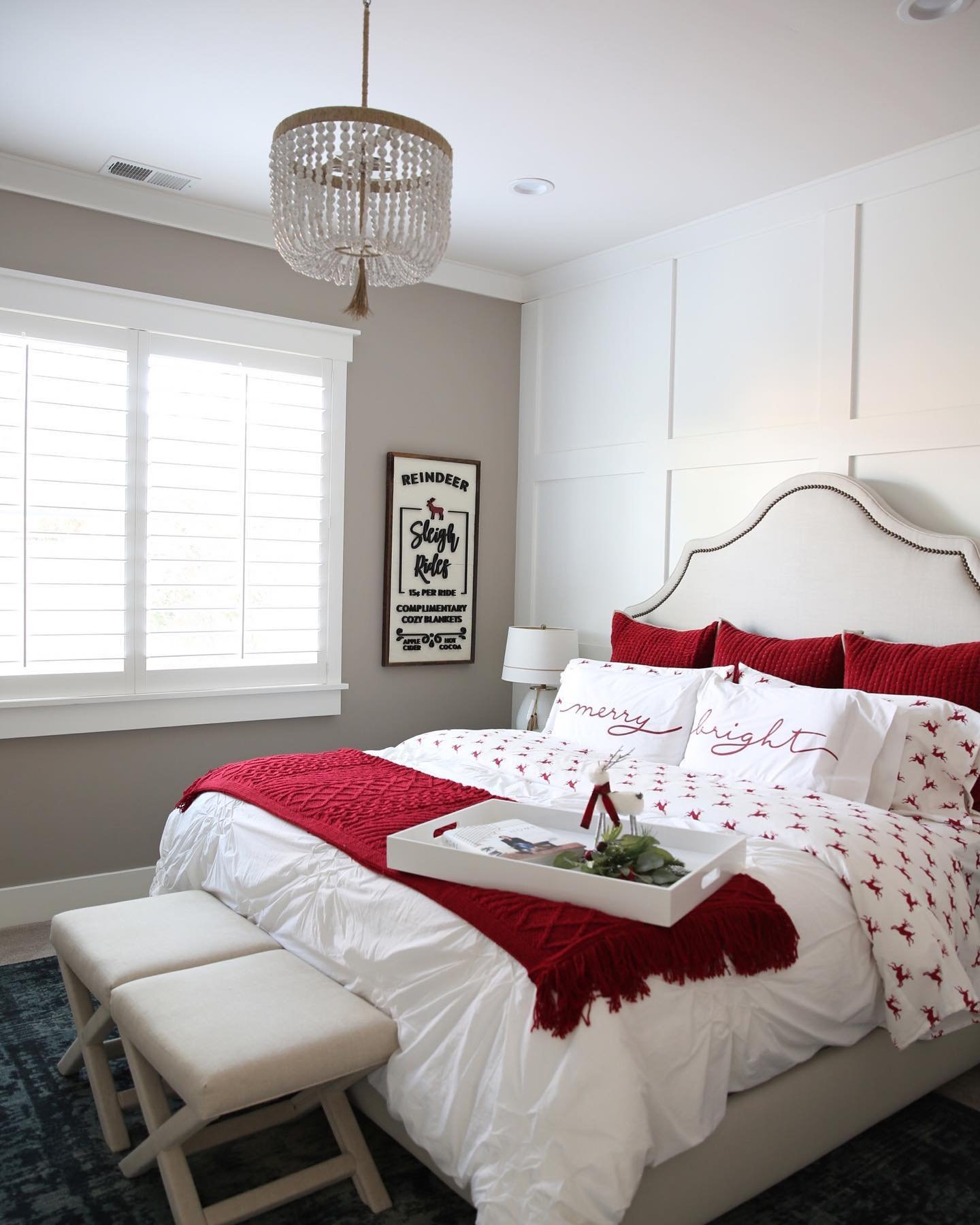 A bright bedroom with a white upholstered headboard, white and board-and-batten accent walls, and a bed dressed in white bedding with a red reindeer pattern and solid red accents.