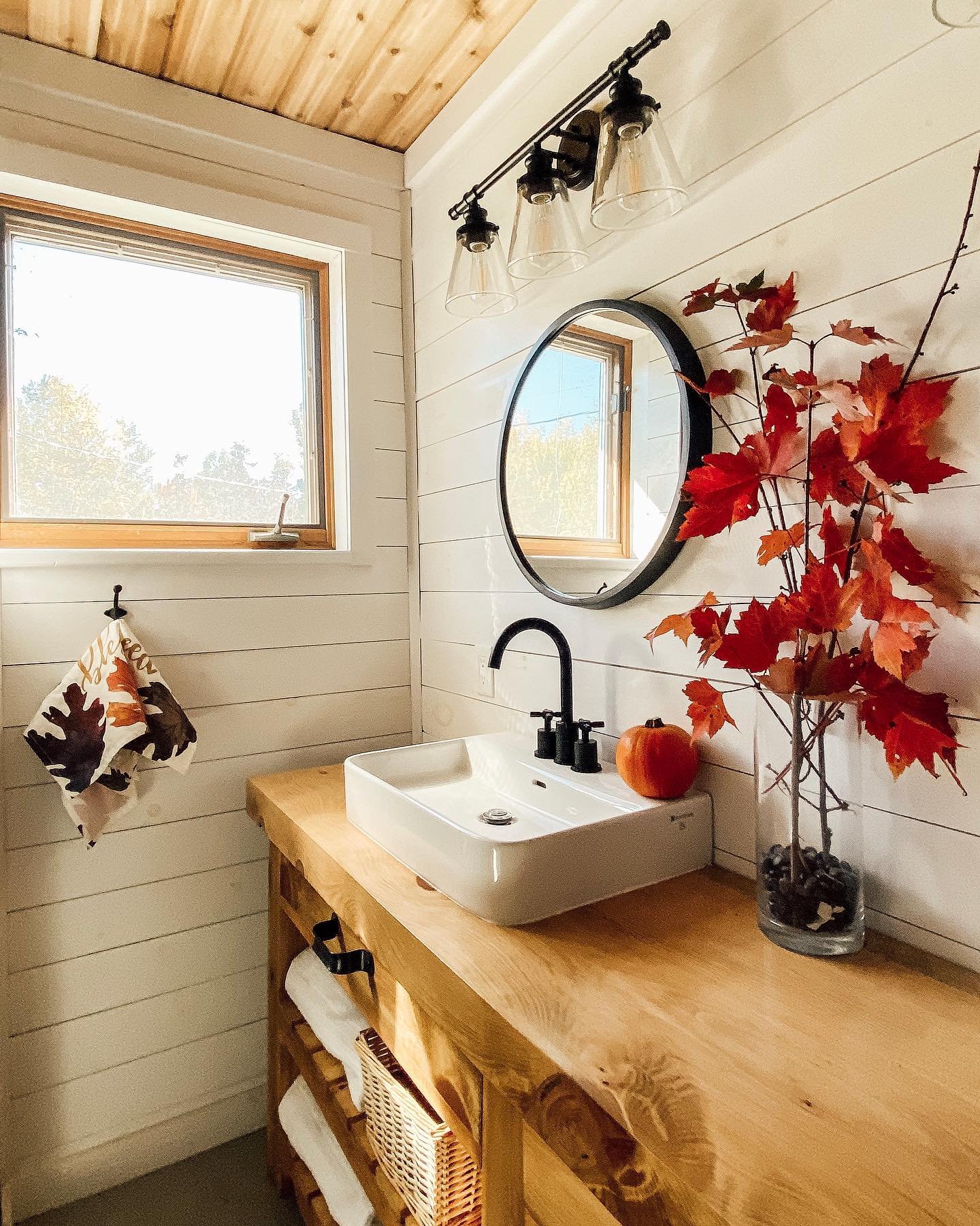 A modern, minimalist bathroom with a wood counter and white shiplap walls, featuring a vase of bright red autumn leaves.