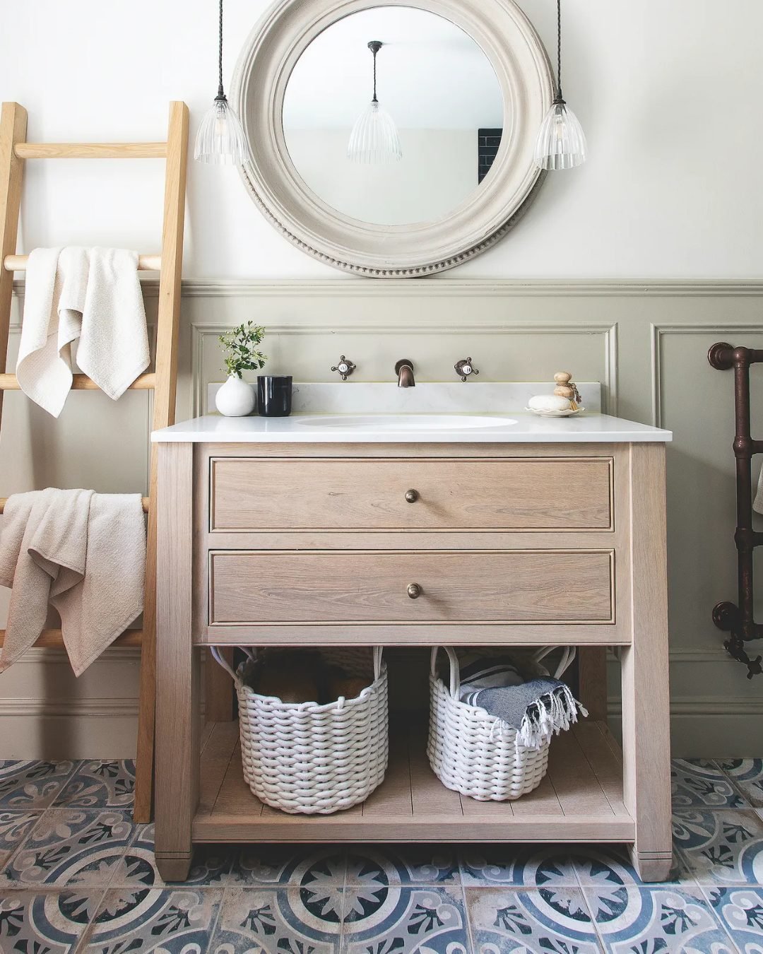 Light wood bathroom vanity featuring two drawers, an open lower shelf containing two large white woven baskets, and a leaning wooden towel ladder next to it.