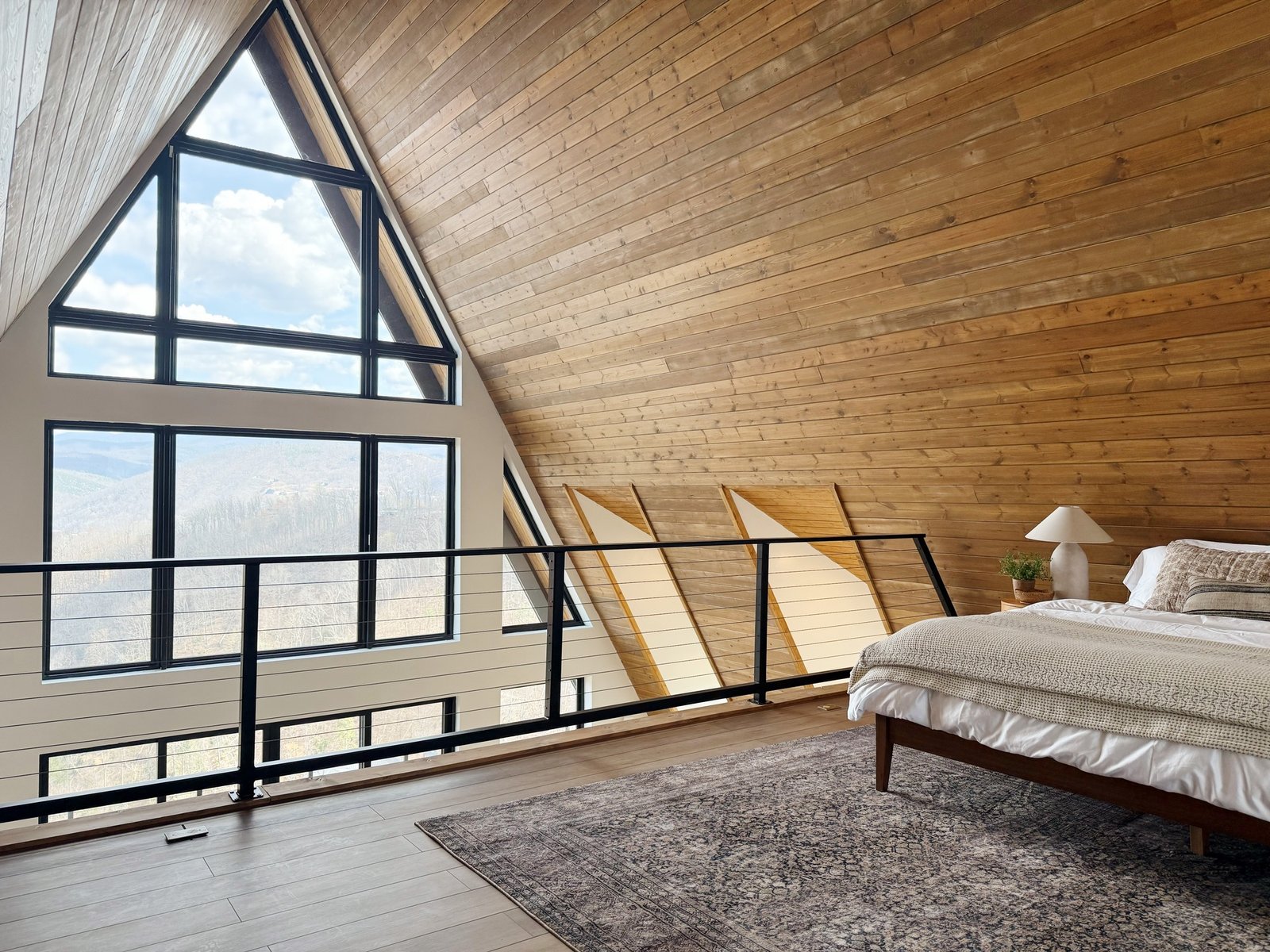 A rustic A-frame loft bedroom with a large triangular window overlooking mountains, a sloped wood plank ceiling, and a bed with neutral bedding.