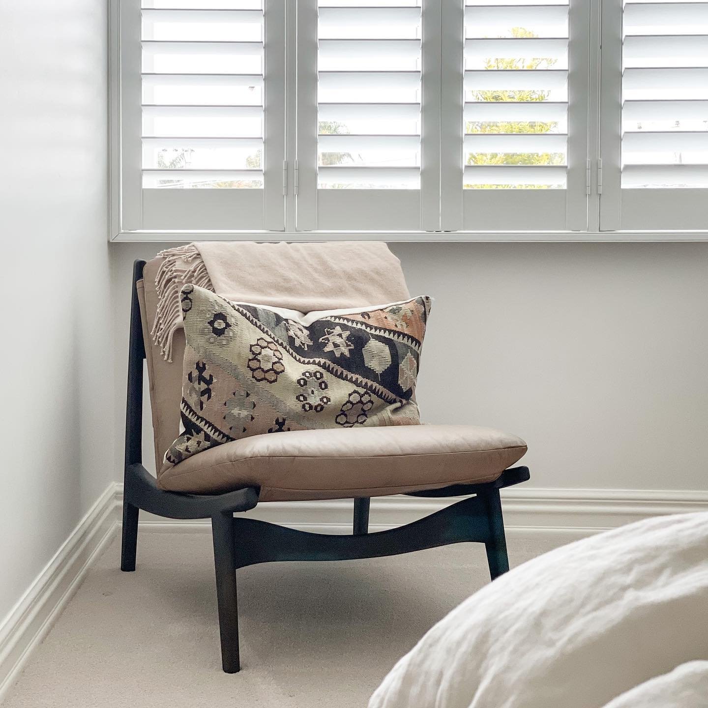 A minimalist slipper chair with a dark wooden frame and a beige leather seat sits in a bedroom corner next to a window.
