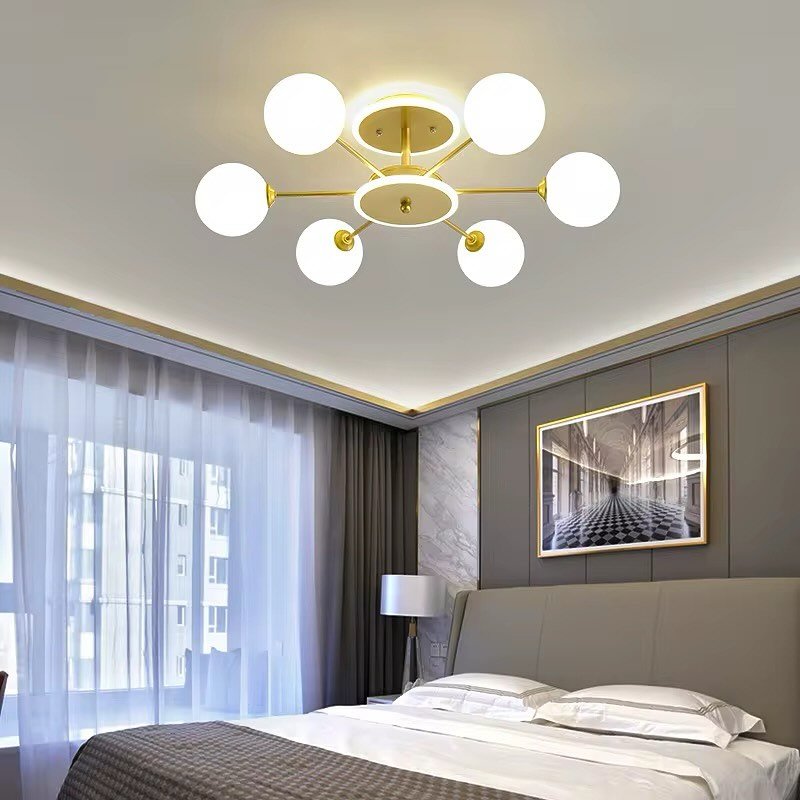 A contemporary semi-flush mount ceiling light with a gold finish and six white globe shades arranged in a starburst pattern hangs over a bed.