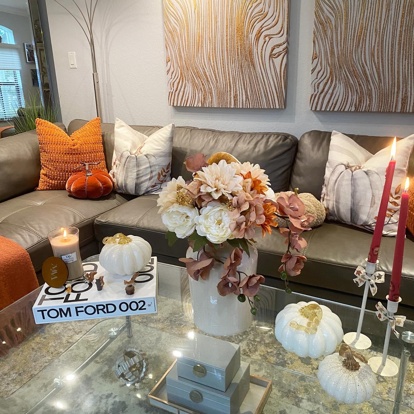 Gray leather sofa with orange and pumpkin-themed pillows, a glass coffee table with white pumpkins and candles, and gold art on the wall.