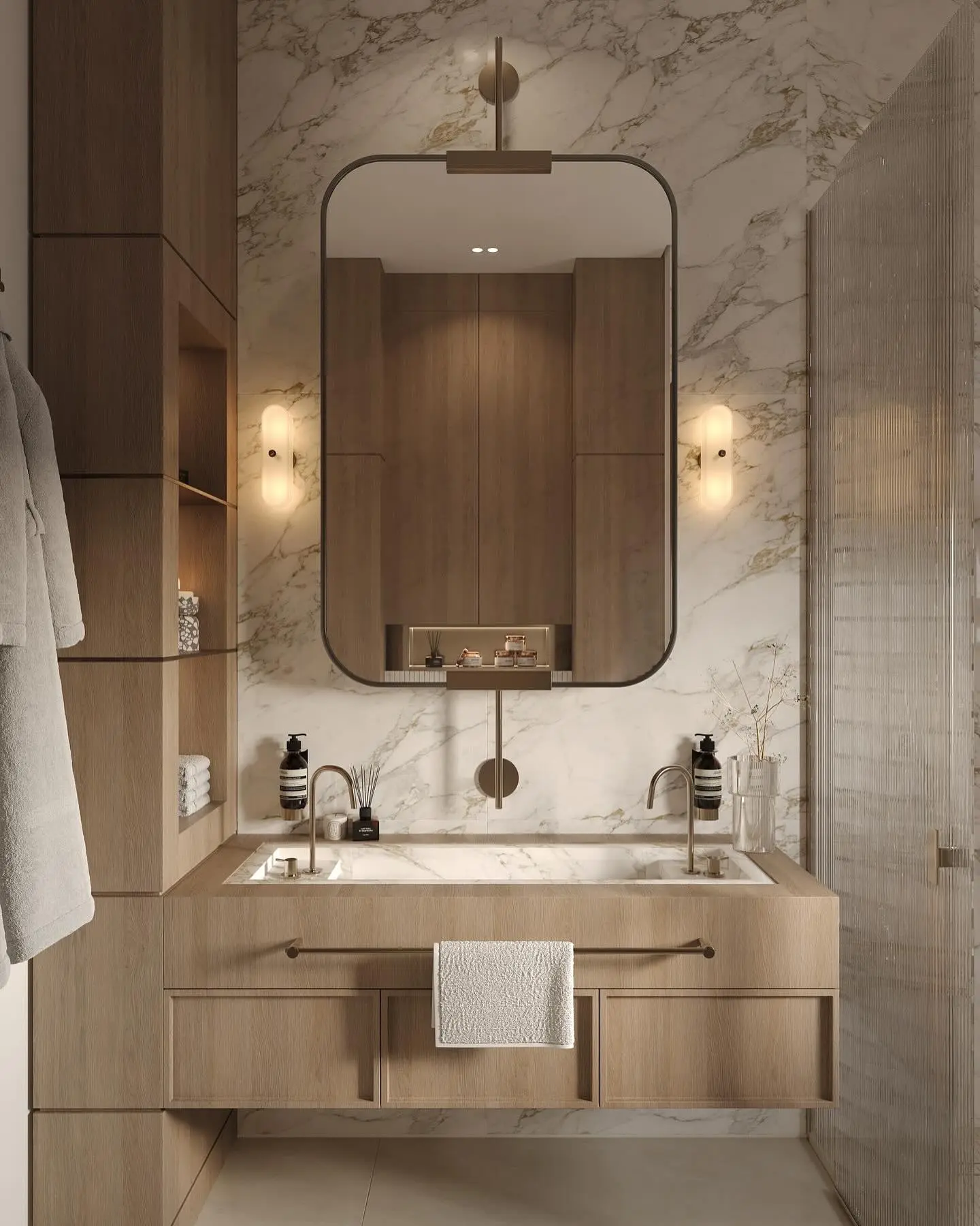 A luxurious double vanity with a light wood base and an integrated white marble countertop and sink. It features two gold faucets, a large black-framed mirror, and two vertical sconces. The vanity is set against a wall of white marble with gold veining.