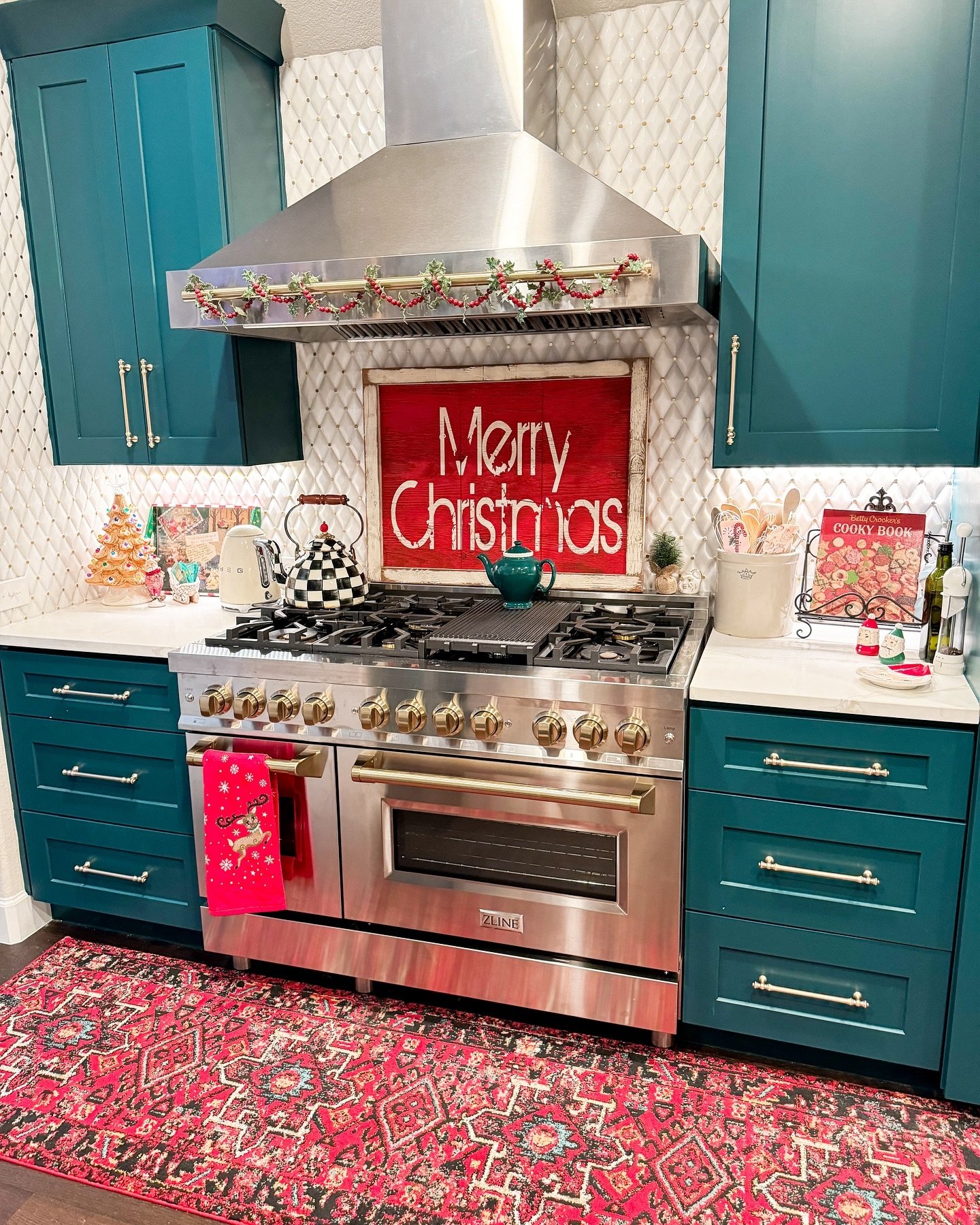 A vibrant, maximalist kitchen featuring striking teal cabinetry, a professional stainless steel range, and a distressed red sign that reads "Merry Christmas" set against a quilted white backsplash.