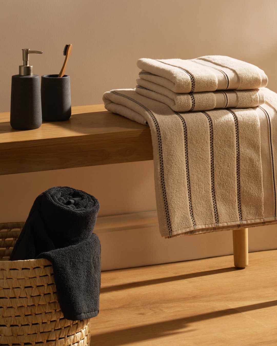 Stack of beige towels with thin black stripes on a wood bench, with a black rolled towel in a wicker basket below.