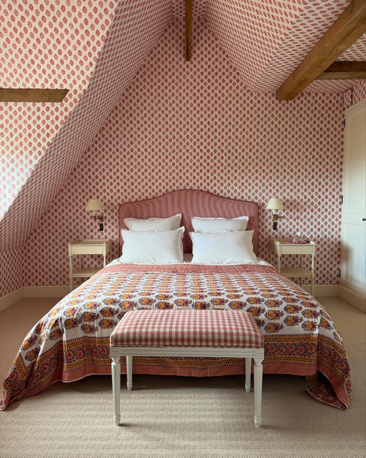 A cozy attic bedroom with a sloped ceiling and walls covered in a red and white patterned wallpaper, a bed with a pink striped headboard, and a floral quilt.