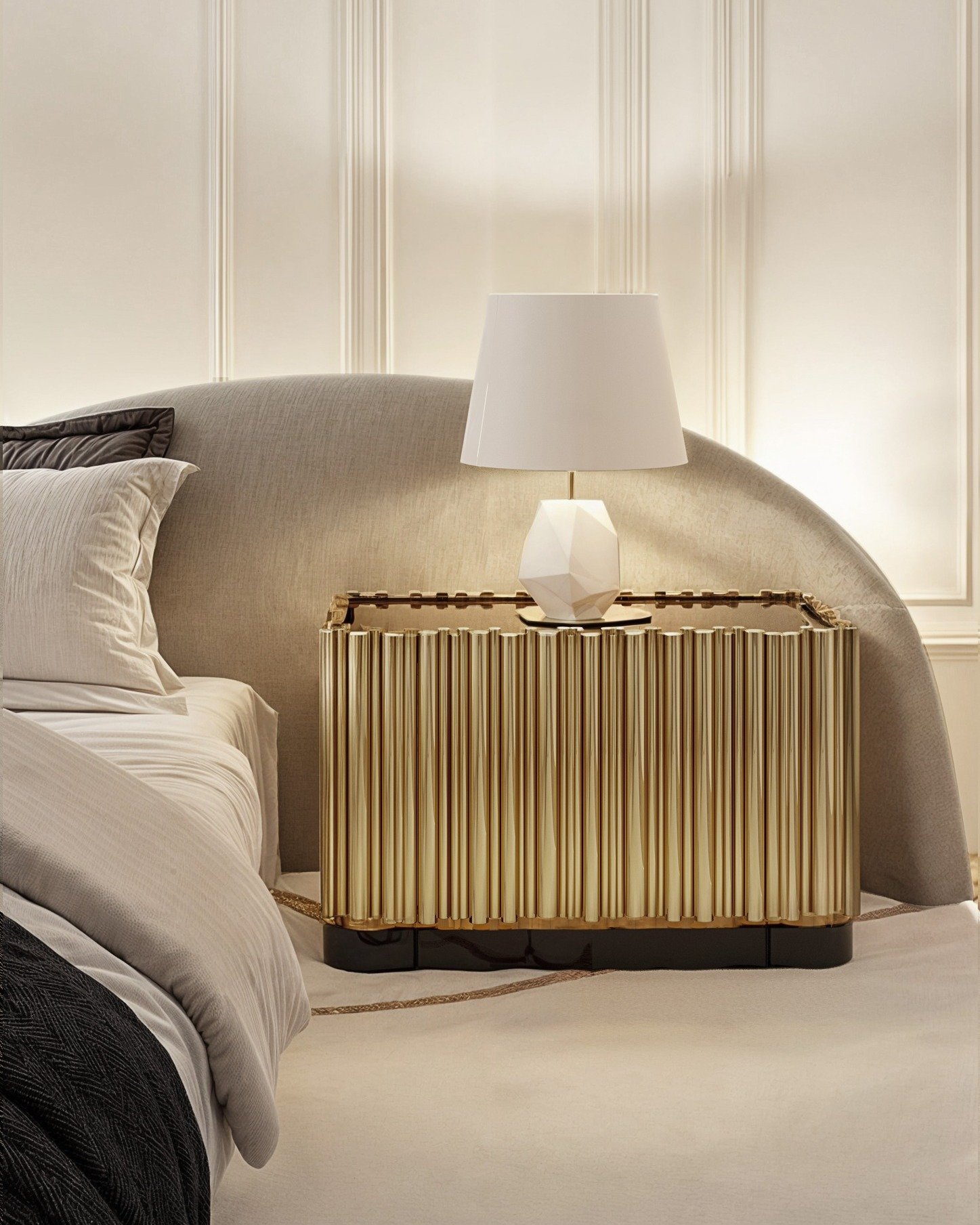 An exquisite, highly ornate nightstand with a metallic gold finish and vertical fluted details. The top features a dark edge and holds a white geometric table lamp.