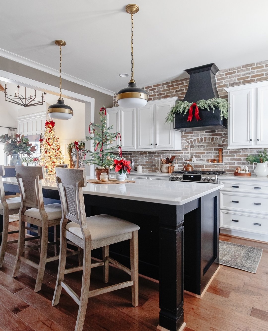 A stylish kitchen with a black island, white shaker cabinets, and an exposed brick backsplash. A dark range hood is adorned with a simple pine garland and a bright red ribbon. A small, sparse Christmas tree sits on the island.