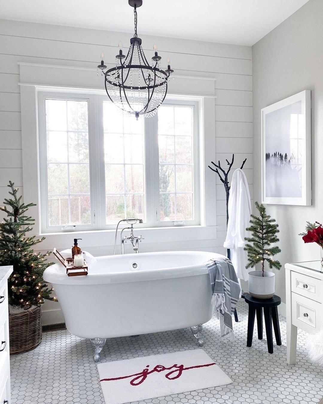 A luxurious white bathroom with a clawfoot tub, two potted Christmas trees flanking the tub, and a red "joy" bath mat.
