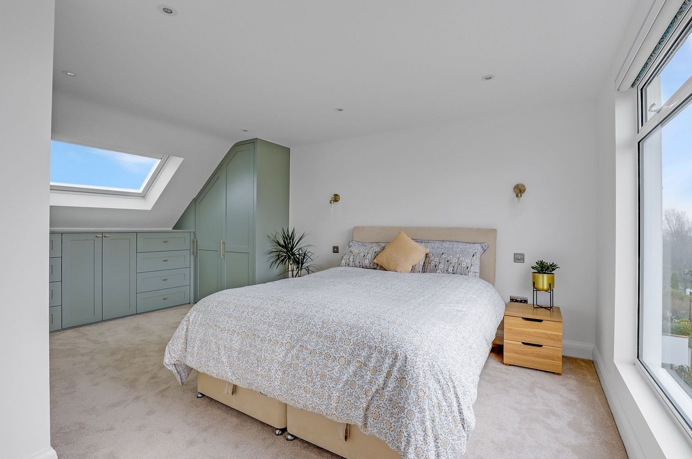 A bright loft bedroom with white walls, a light bed, and custom-built sage green cabinets and a wardrobe built into the sloped wall.