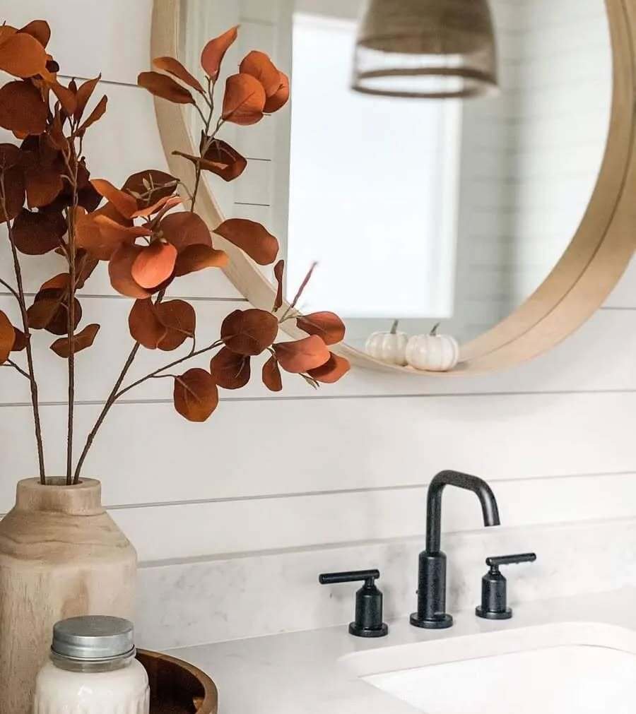 A modern bathroom with a light wood-framed mirror and a vase of burnt orange eucalyptus branches on the counter.