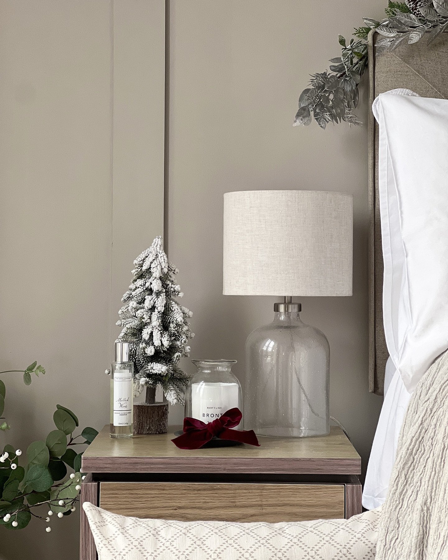 A clear glass table lamp with a linen shade sits on a two-toned wooden nightstand in a neutral-colored bedroom.