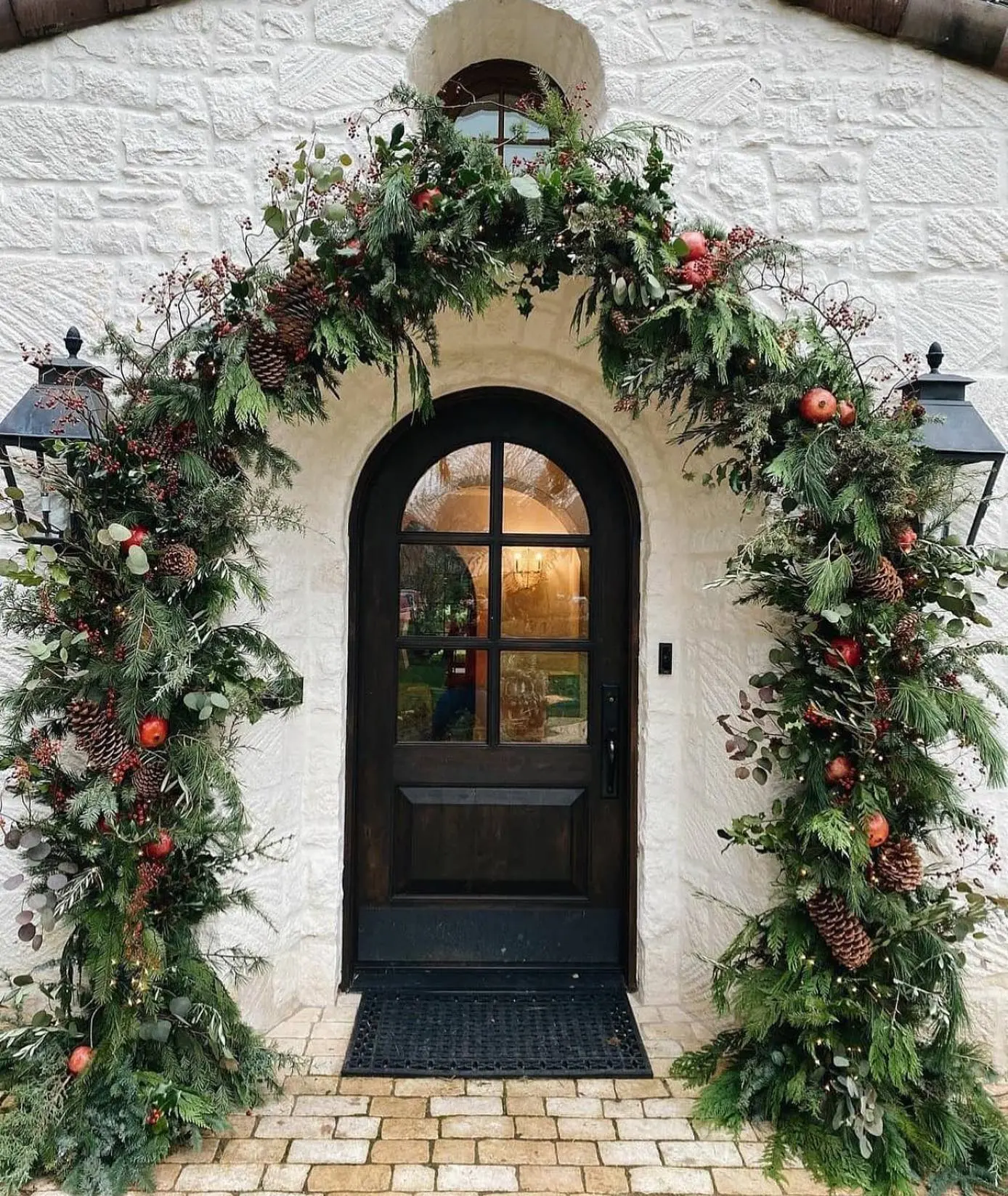 A dramatic Christmas porch with a black arched door set against a white stone wall, framed by an incredibly full, oversized garland decorated with pinecones, red berries, apples, and eucalyptus.