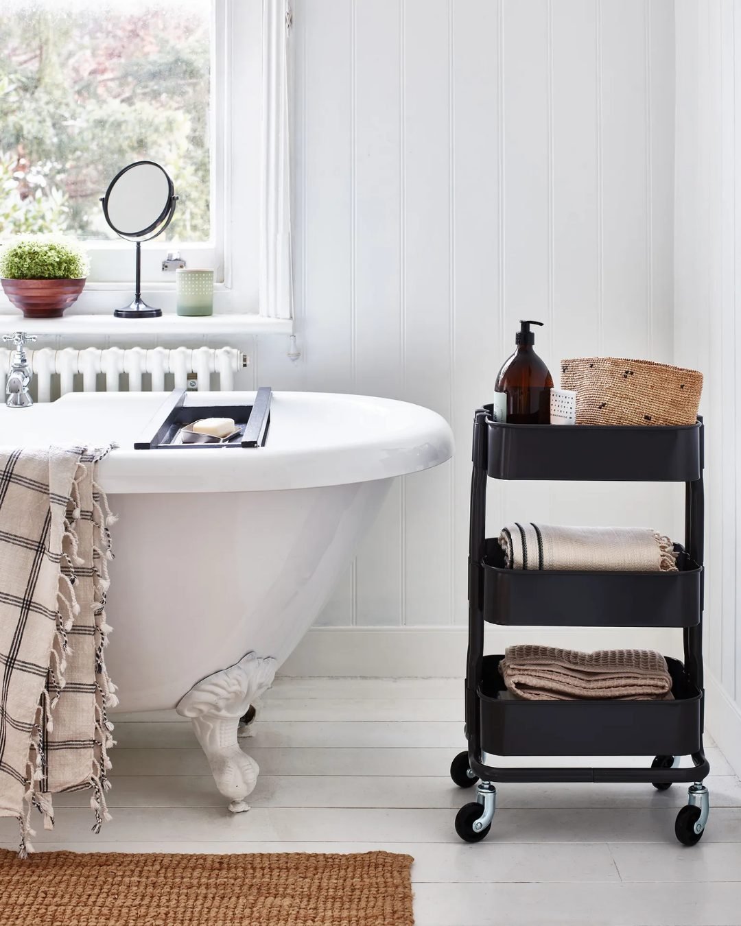 Clawfoot bathtub next to a window and a black metal three-tier rolling utility cart used to store towels, a dispenser bottle, and a small woven basket.