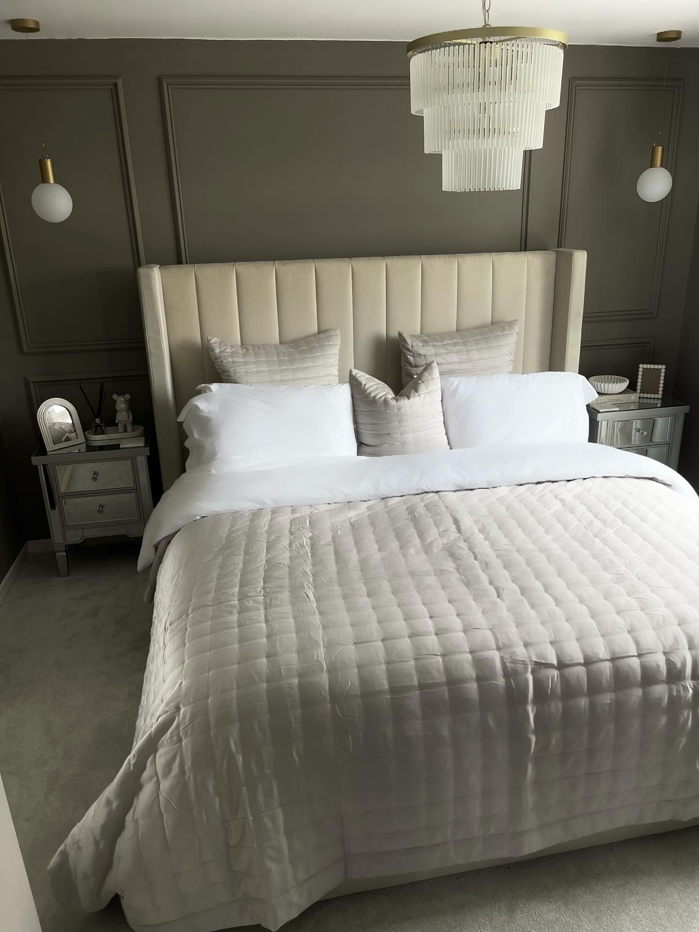 A multi-tiered chandelier with layers of clear vertical glass rods hangs over a bed in a glamorous, neutral-toned bedroom.