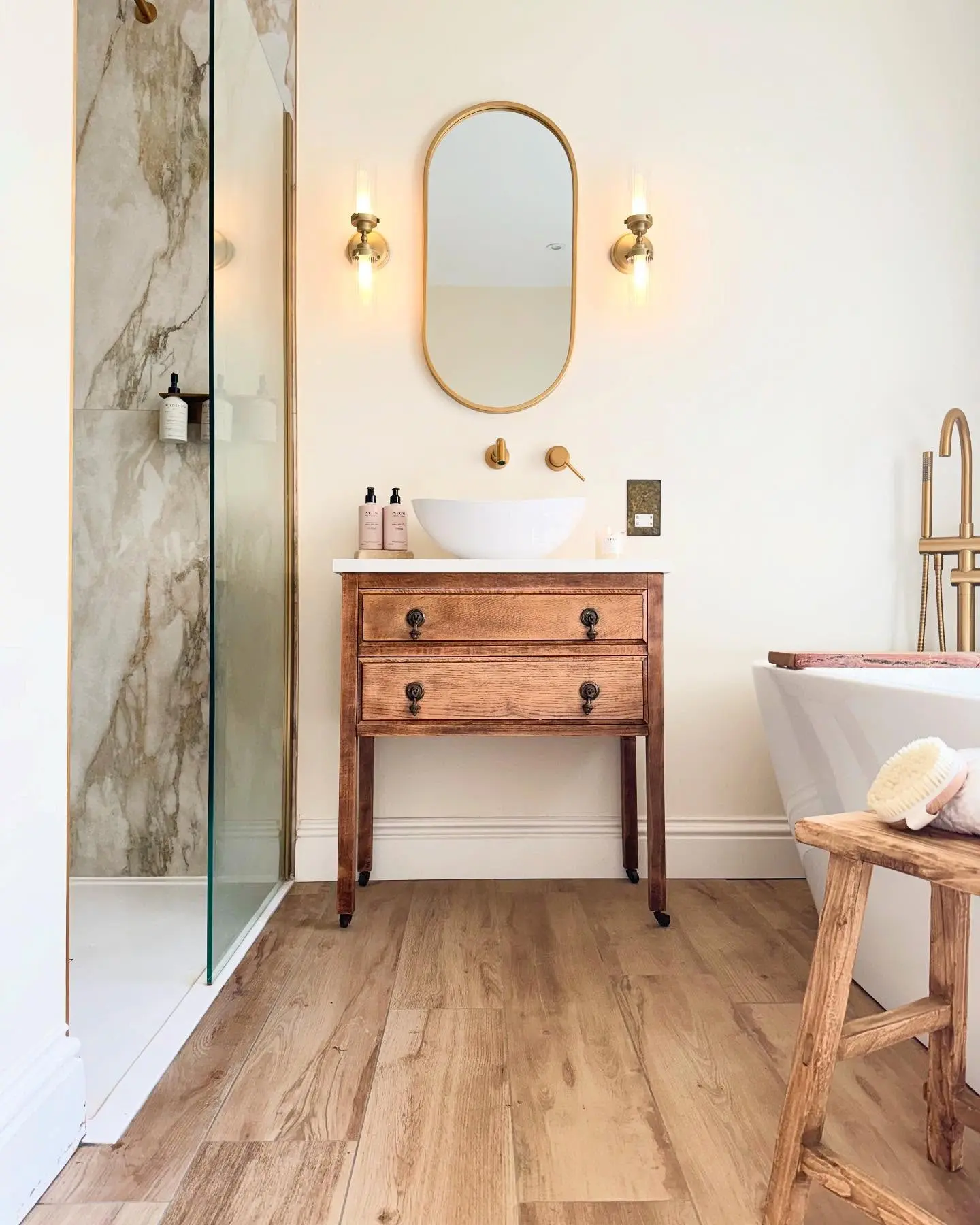 A small bathroom vanity that looks like a vintage wooden console table with two drawers and black drop handles. It has a white countertop and a vessel sink, paired with a gold-framed pill-shaped mirror, wall-mounted gold faucet, and two wall sconces.