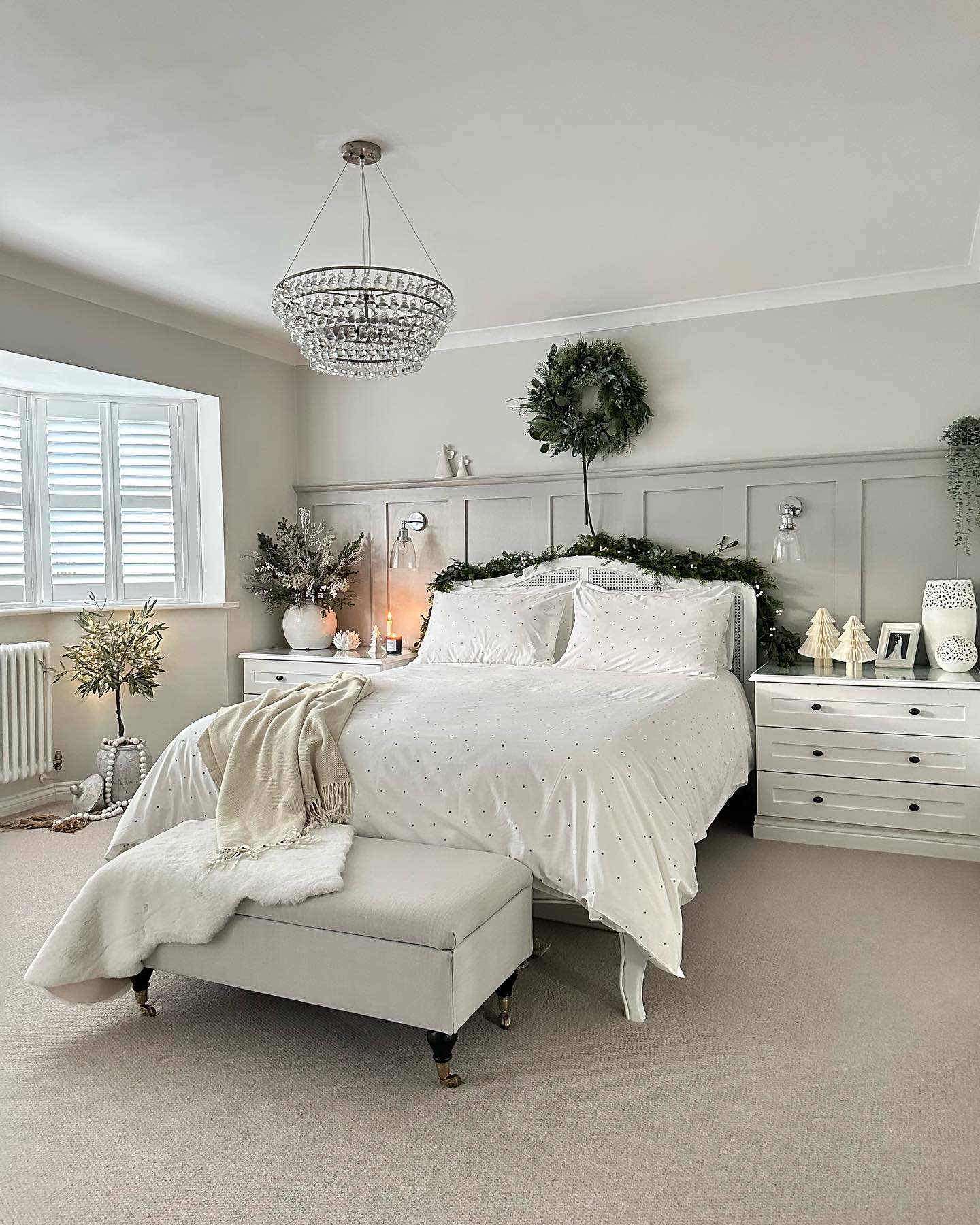 A bright, elegant bedroom with gray paneling, a crystal chandelier, all-white bedding with a subtle pattern, and greenery including a wreath and garland draped over the headboard.
