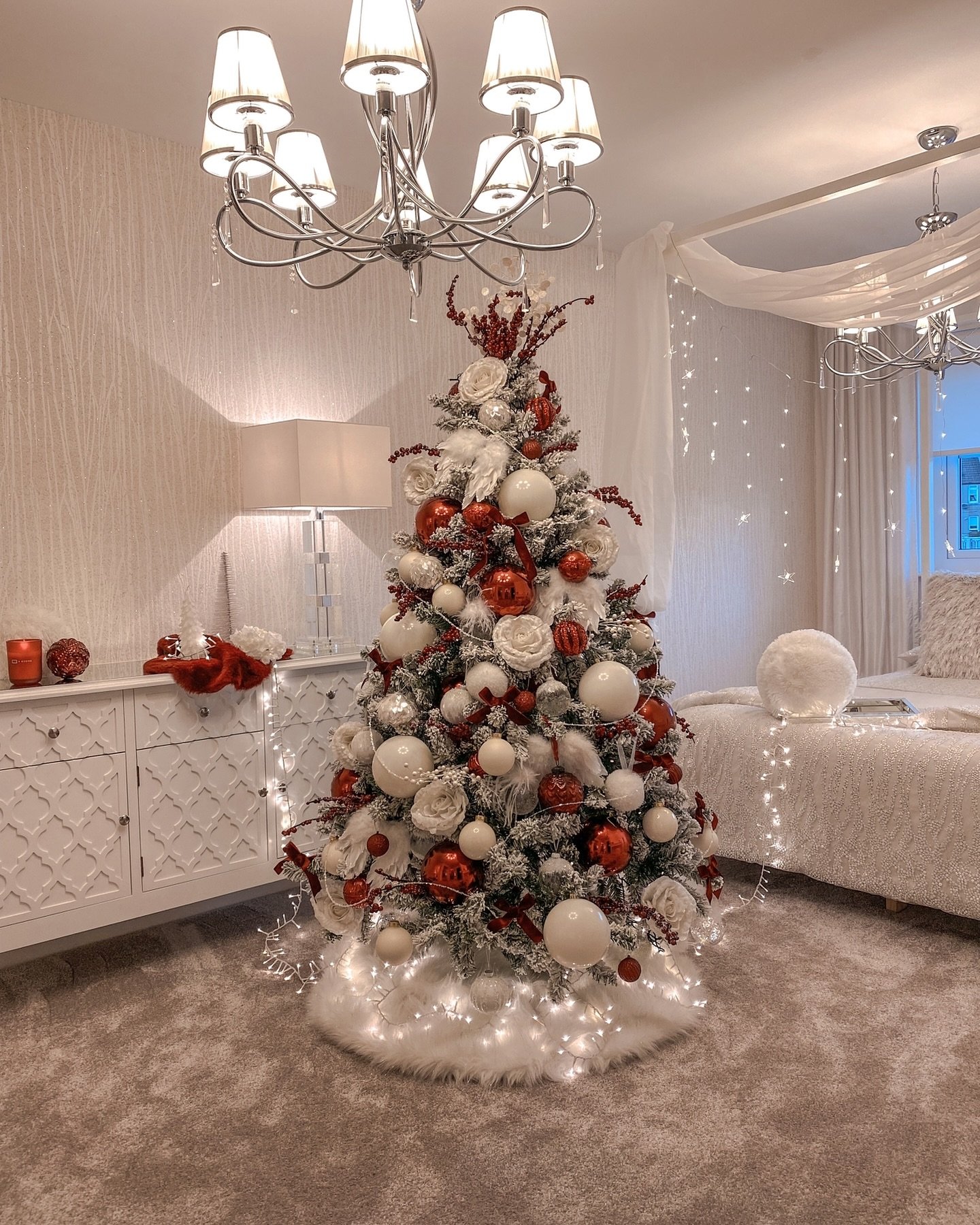 A dense, heavily flocked Christmas tree decorated with bold red and large white baubles, and oversized white floral accents in a bright bedroom setting.