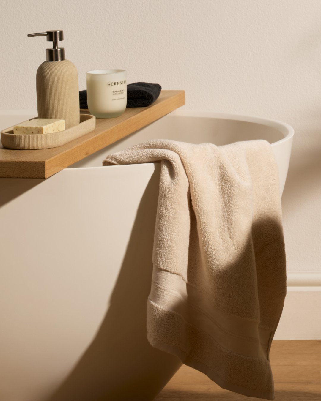 Cream bath towel draped over the side of a white bathtub with a wooden tray holding beige bath accessories.