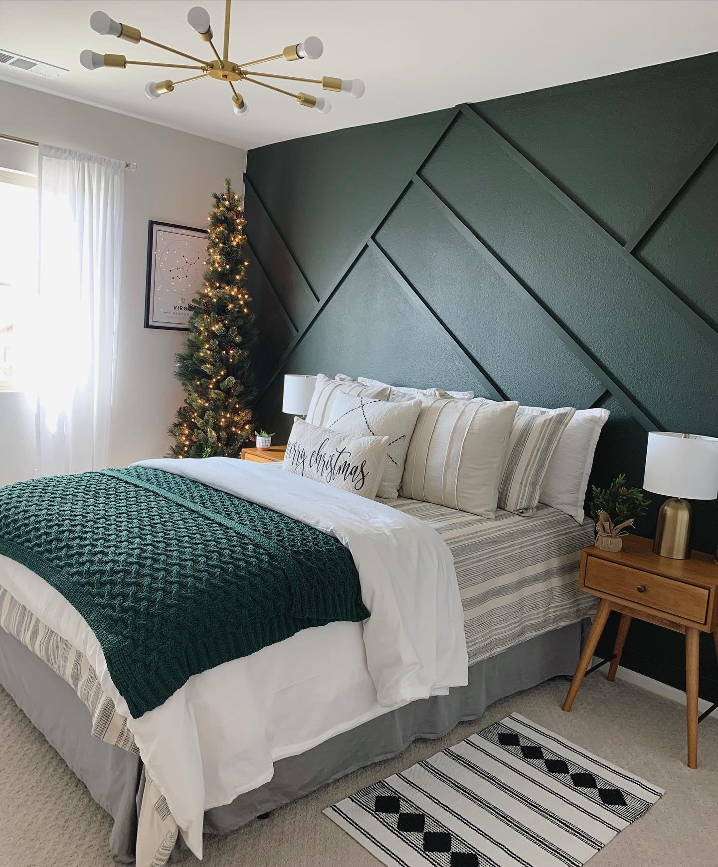 A contemporary bedroom with a striking dark emerald green geometric accent wall, featuring a bed with a chunky cable-knit green throw blanket and a slim, illuminated Christmas tree in the corner.