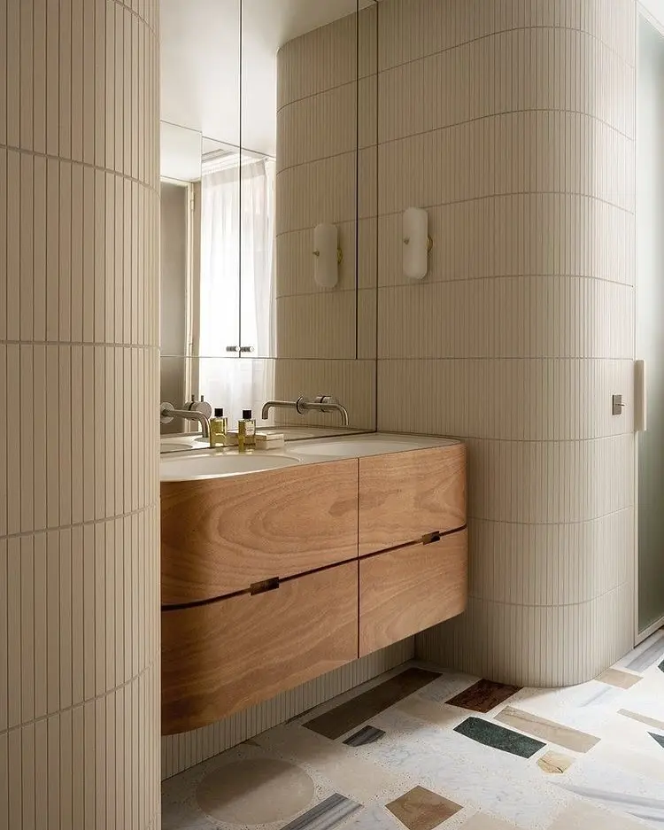 A floating, light wood bathroom vanity with curved edges and an integrated countertop and sink. It's paired with a seamless mirror that covers the wall, two modern vertical wall lights, and a floor tiled with various geometric stone shapes.