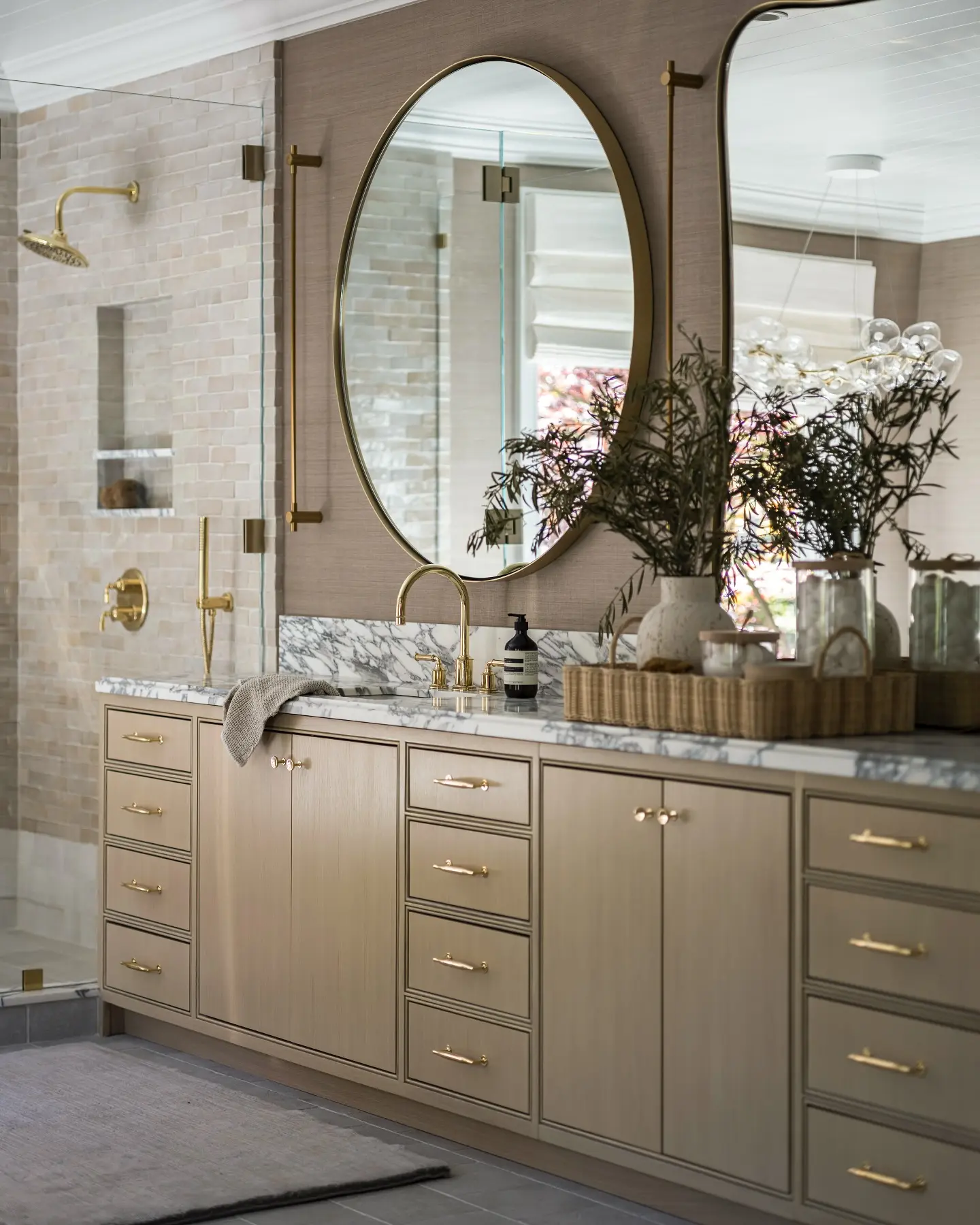 An elegant bathroom with a large oval mirror with a thin brass frame over a light wood and marble double vanity.