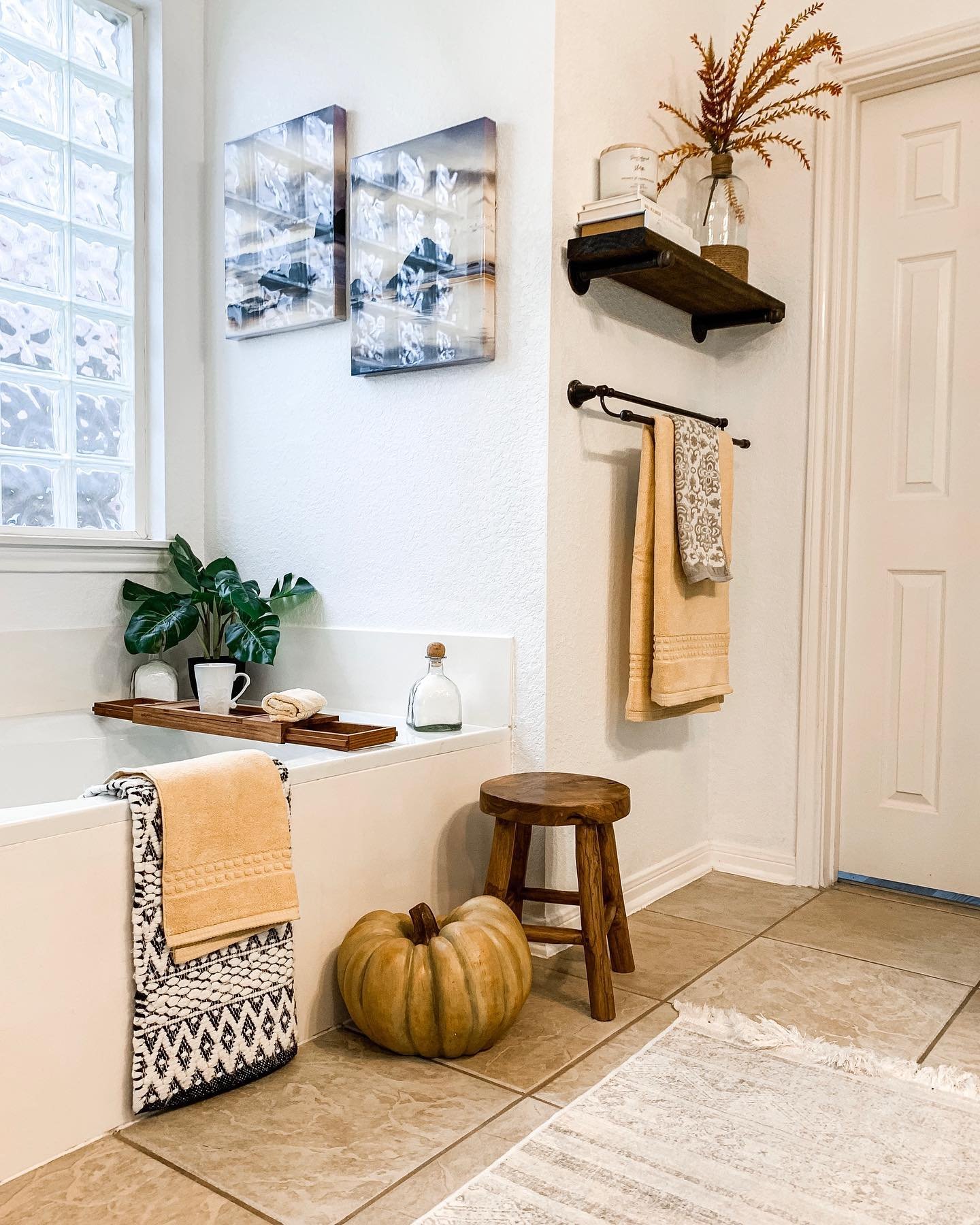 A modern white bathroom with a tub caddy, a large decorative pumpkin on the floor, and a floating shelf with a vase of dry branches.